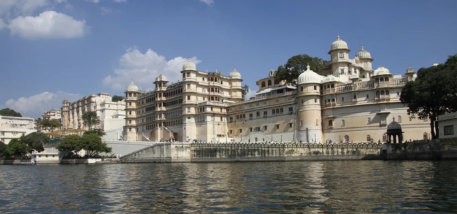 Der cremefarbene City Palace von Udaipur erhebt sich am Ufer des Pichola-Sees unter blauem Himmel mit Wolken