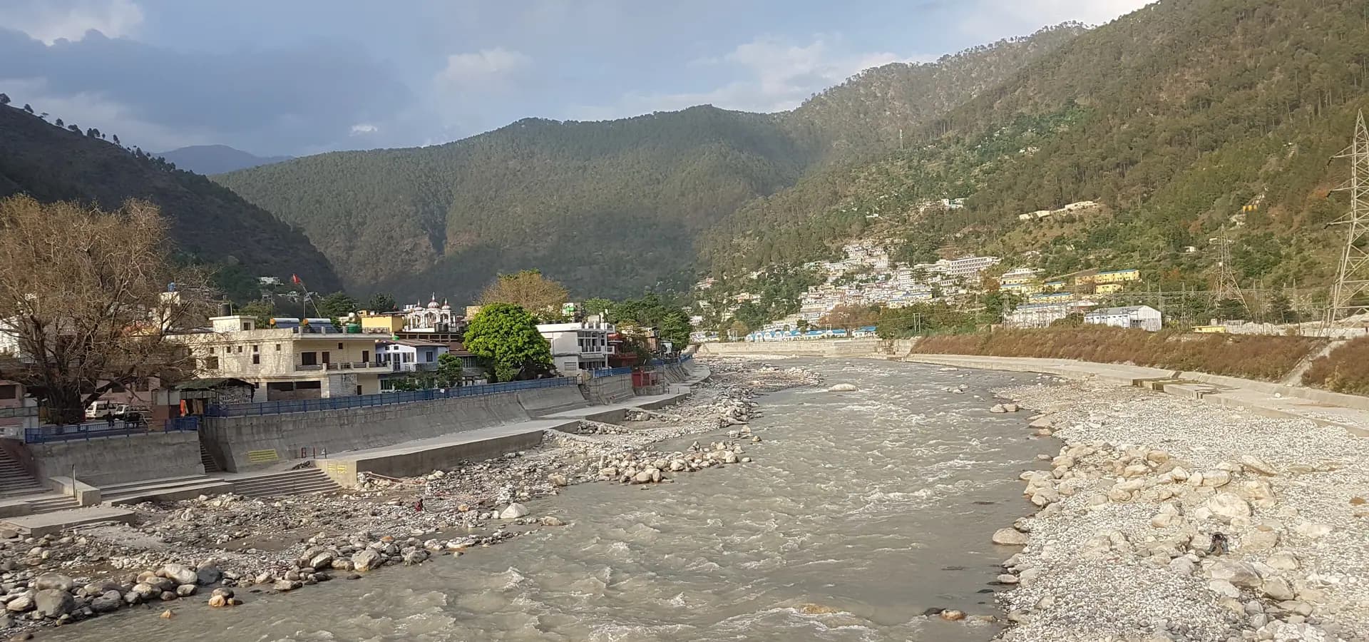O rio Bhagirathi correndo sobre pedras em Uttarkashi, casas nas margens e colinas florestadas sob céu nublado