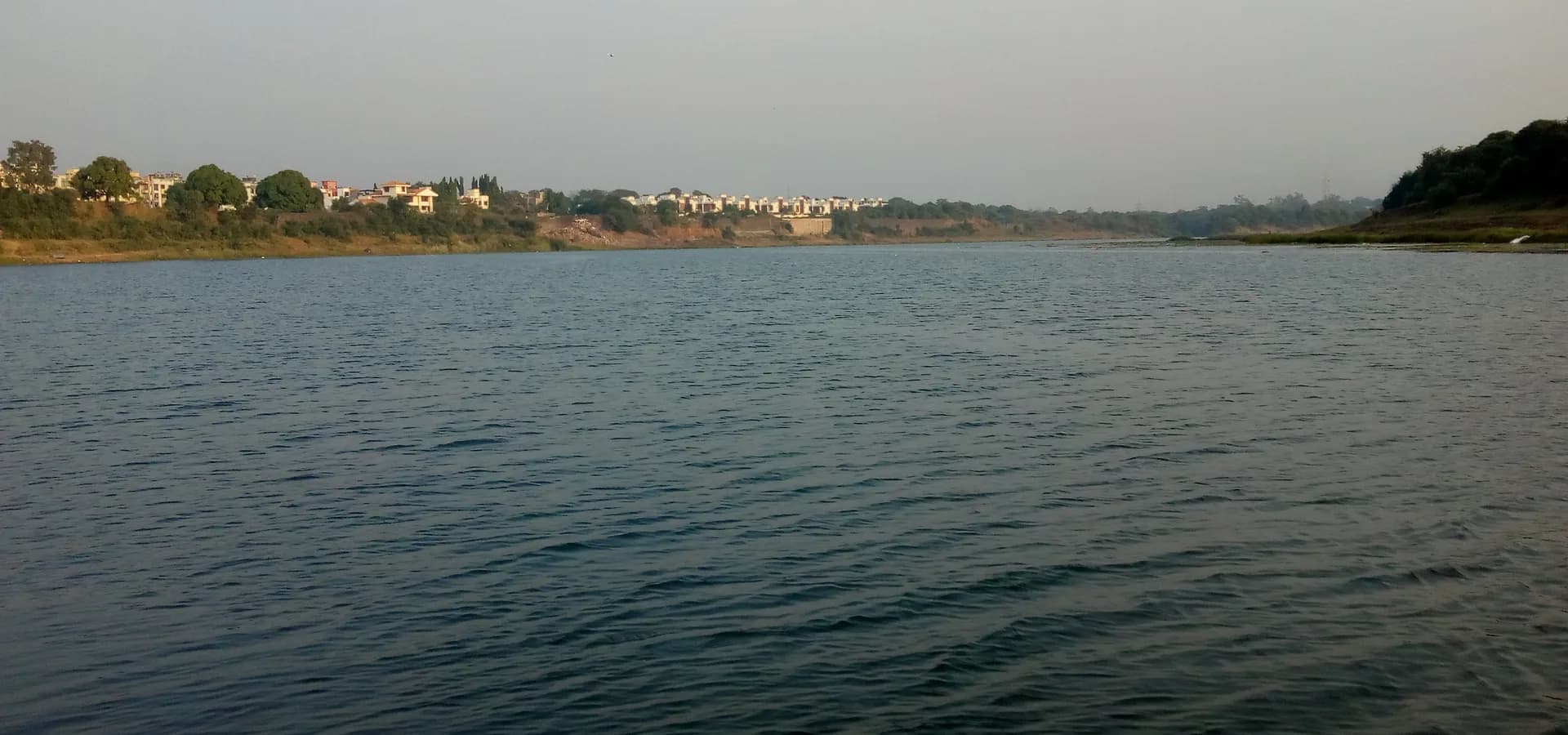 वापी के पास लवाछा में शांत, चौड़ी दमन गंगा नदी, दूर के किनारे पेड़ और घरों की कतार