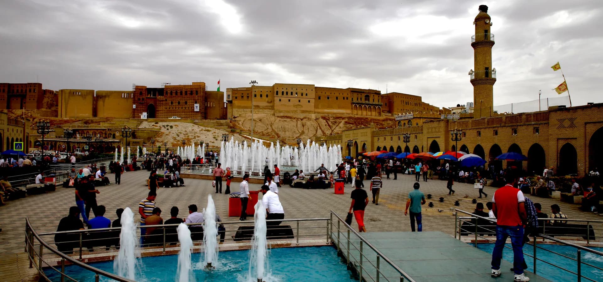 Los largos muros de la ciudadela de Erbil sobre una plaza de fuentes, paseantes y arcadas del bazar bajo un cielo nublado