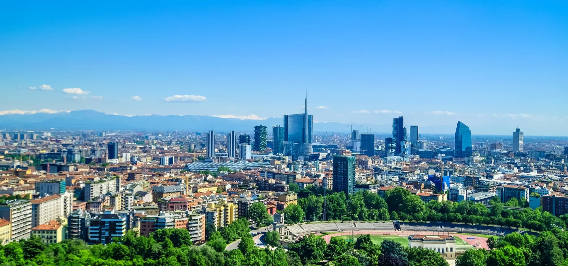 Skyline von Mailand über den grünen Baumkronen des Parco Sempione, Türme von Porta Nuova ragen auf, die Alpen schwach am Horizont