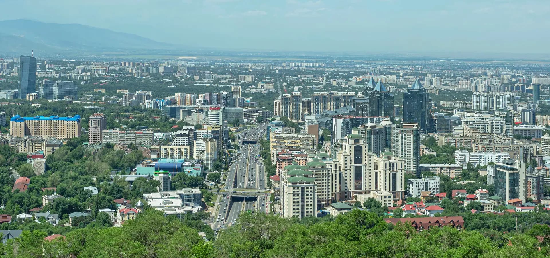 Amplia vista de Almaty desde la colina Kök Töbe, con árboles verdes al frente y torres que se extienden hasta la llanura brumosa