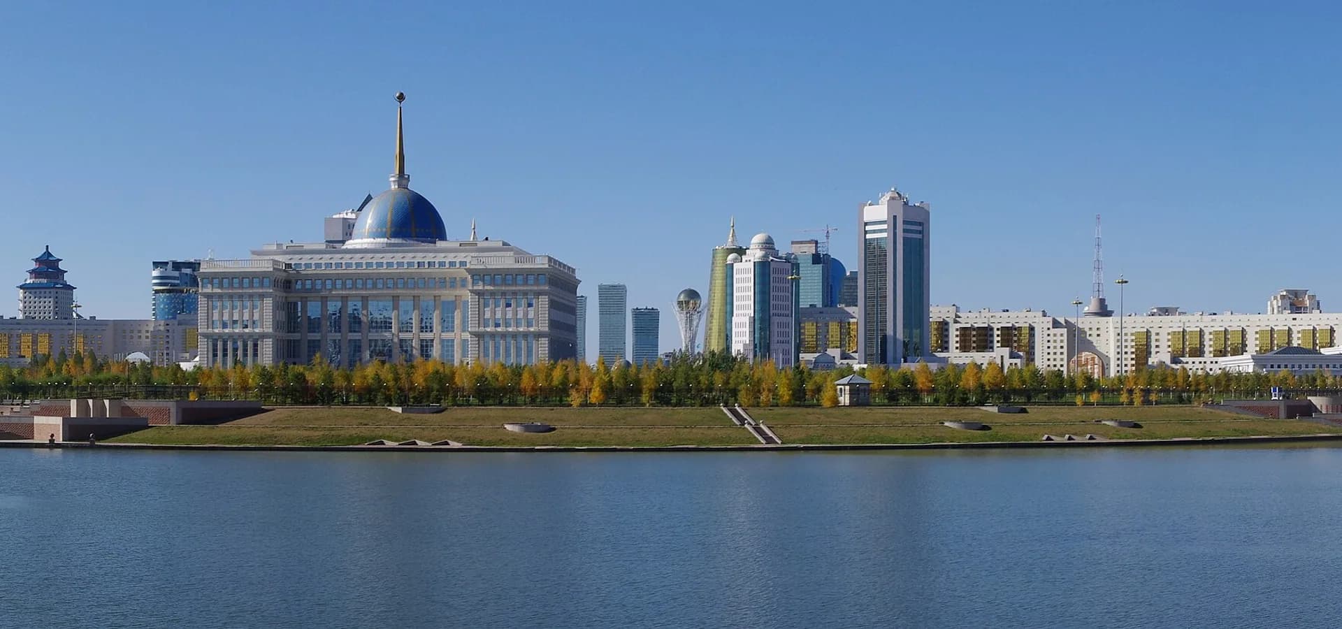 Panorama de la Margen Izquierda de Astana al otro lado del tranquilo río Ishim, con un palacio de cúpula azul, torres y árboles otoñales bajo un cielo despejado