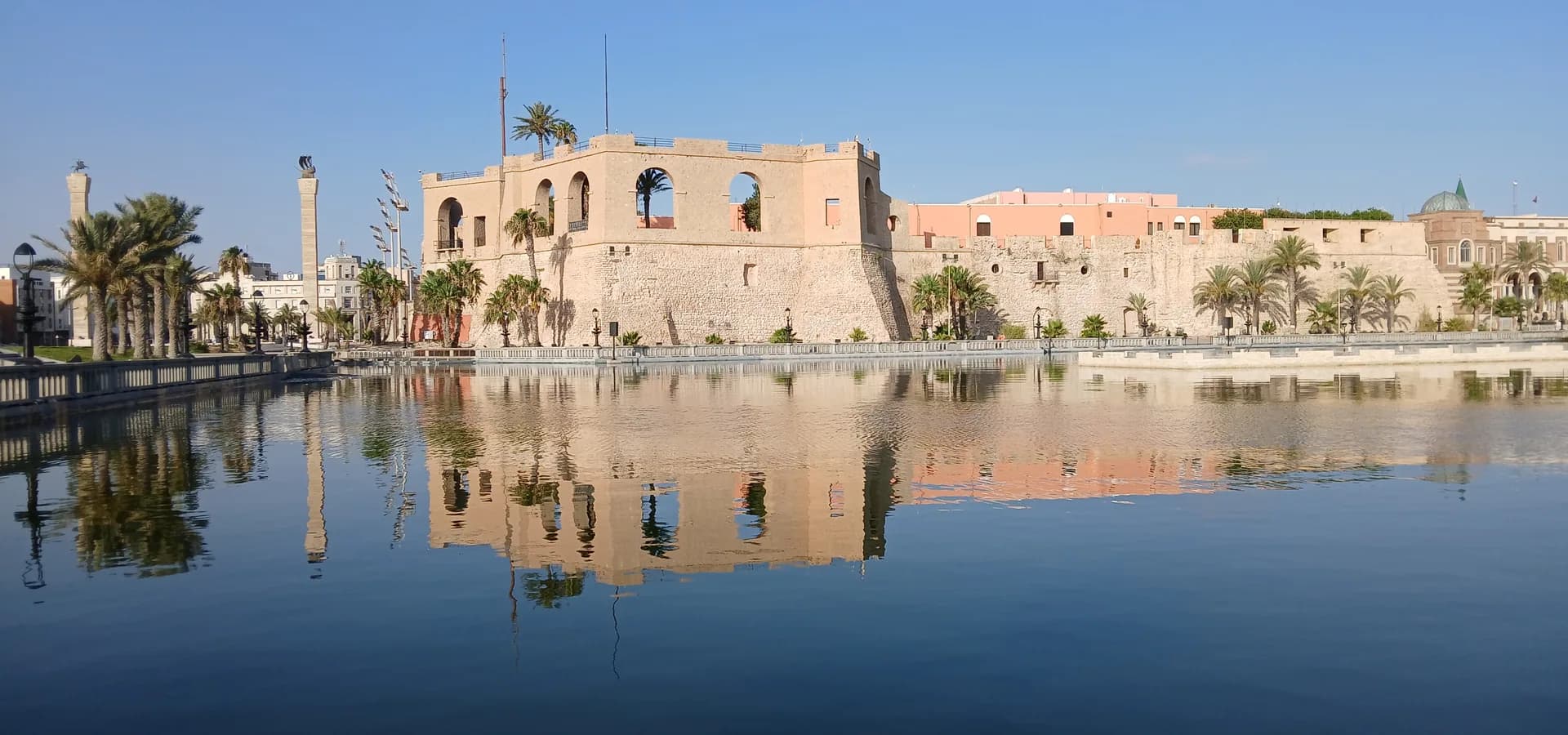 Die sandfarbene Rote Burg von Tripolis mit ihren Palmen, gespiegelt im ruhigen Wasser unter klarem blauem Himmel