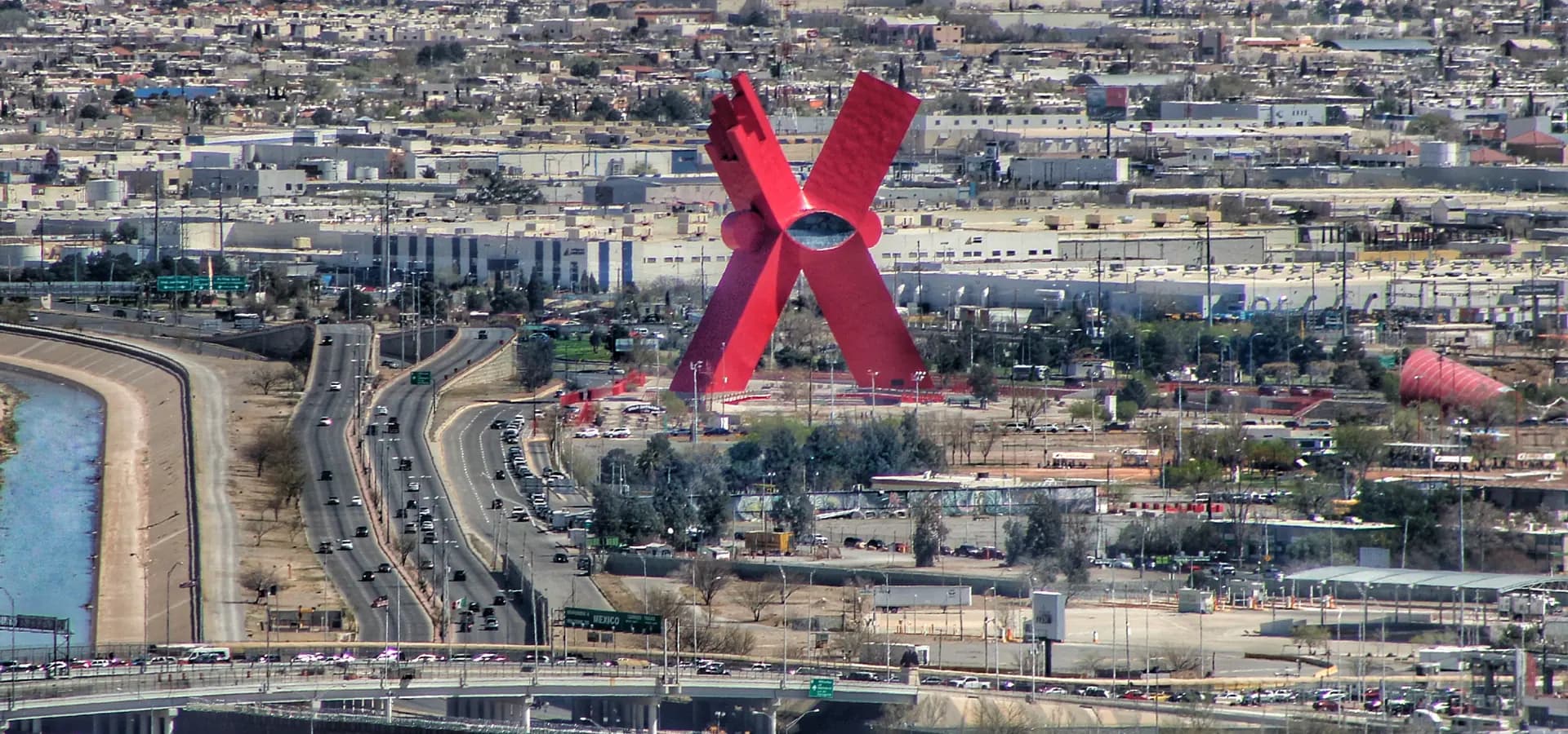 Escultura de um X vermelho gigante sobre a cidade plana de Ciudad Juárez, com o canal do Rio Grande à esquerda