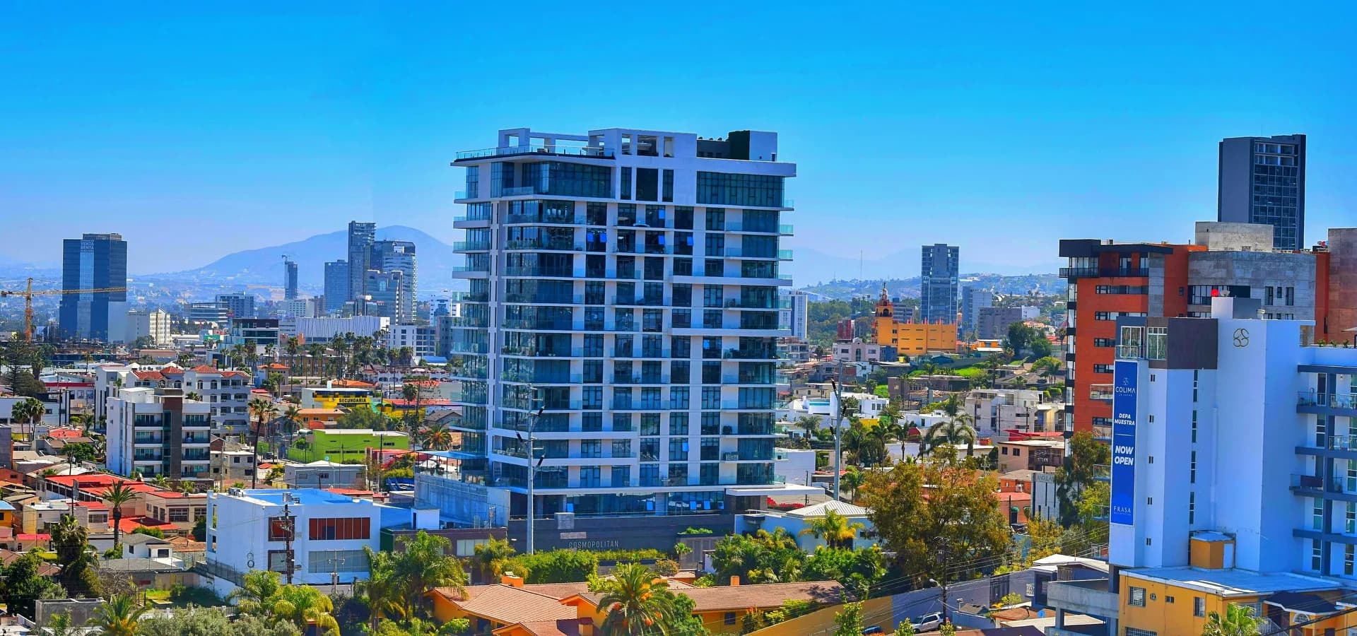 Skyline von Tijuana mit Glashochhäusern, Palmen und niedrigen Häusern vor dunstigen Hügeln unter klarem blauem Himmel