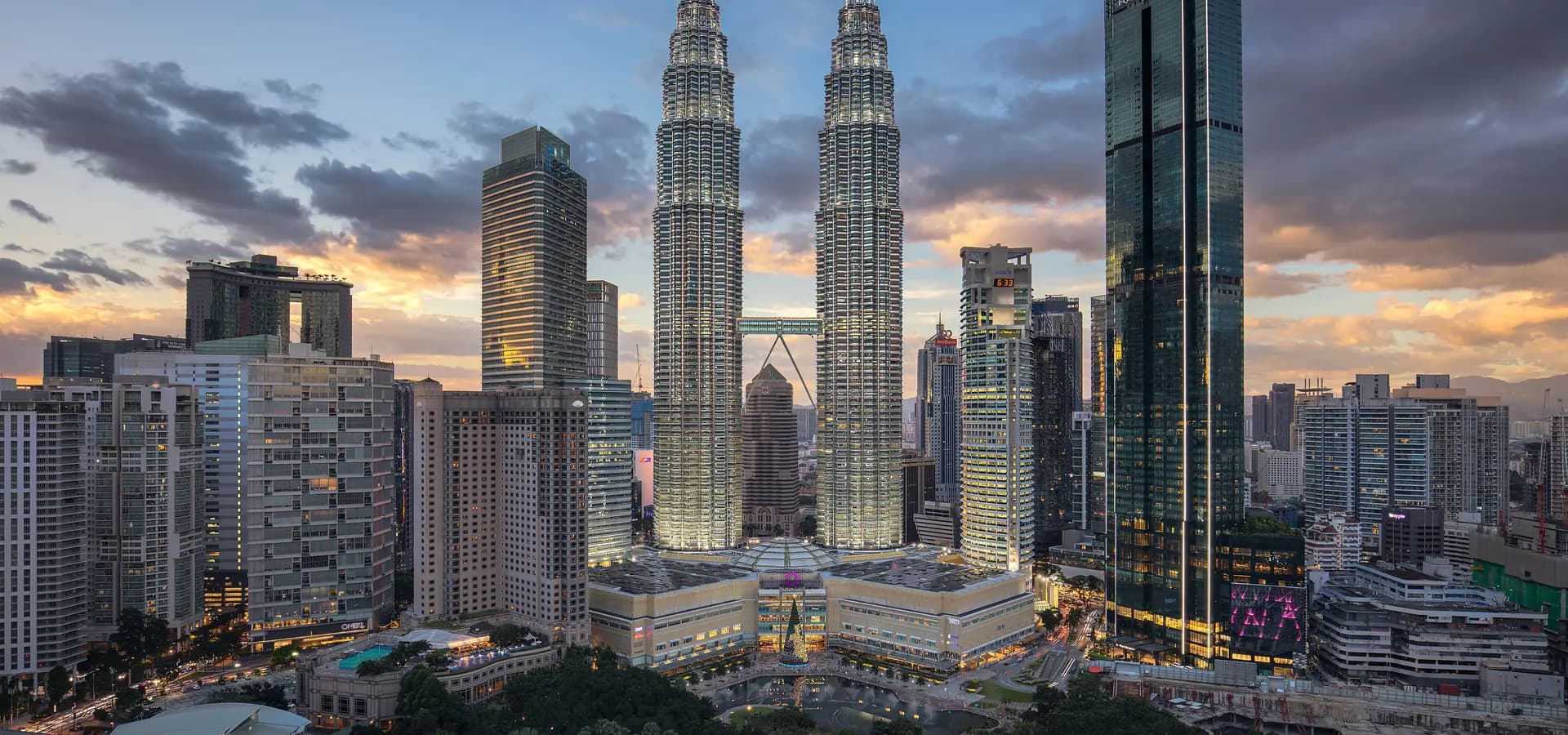 Horizonte de Kuala Lumpur al atardecer, con las torres gemelas iluminadas sobre los árboles del parque KLCC bajo un cielo naranja