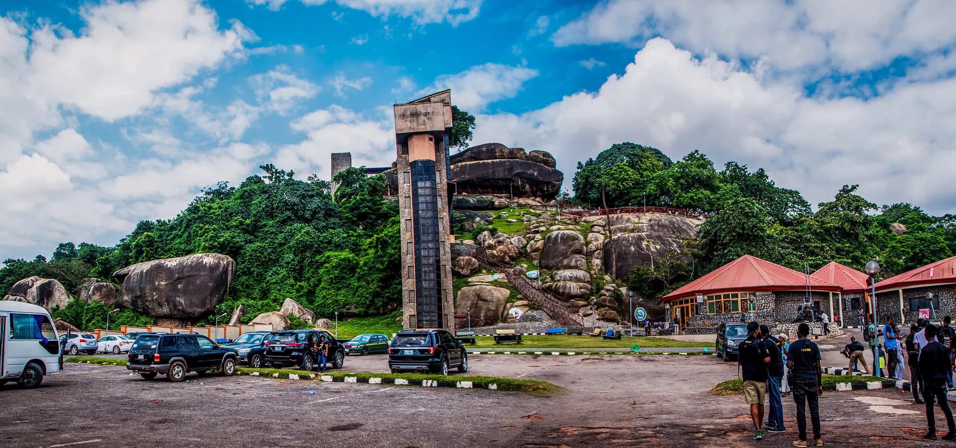 Weite Ansicht des Olumo Rock in Abeokuta, Granitfelsen und grüne Bäume rund um einen Aufzugsturm unter wolkigem blauem Himmel