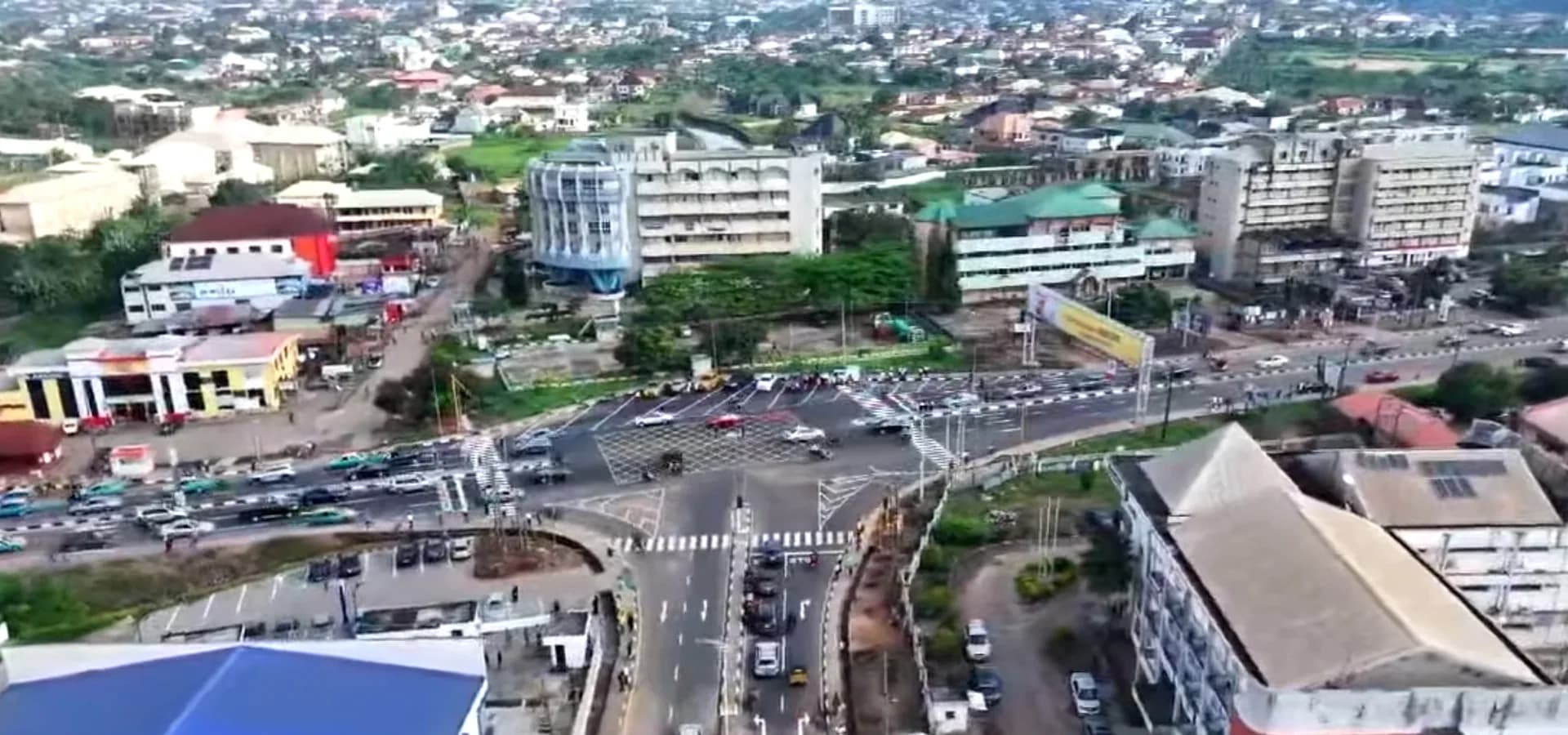 Vista aérea del centro de Akure, un concurrido cruce de calles con edificios de oficinas y techos que llegan hasta colinas verdes
