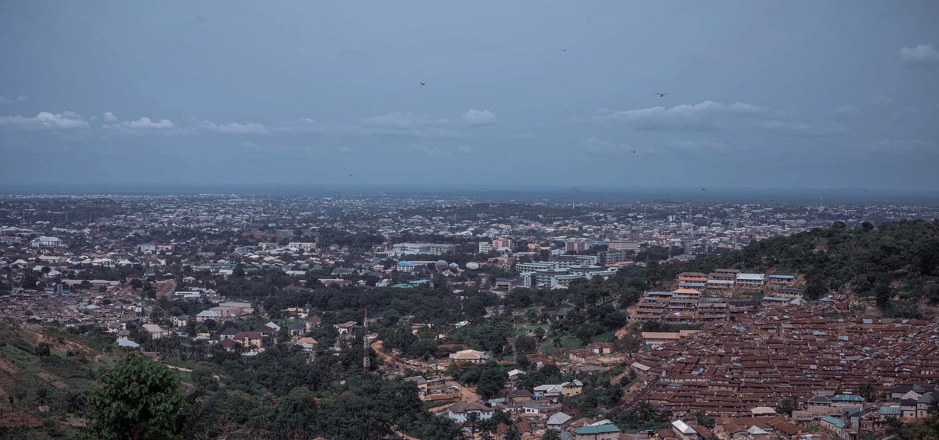 Blick von den Hügeln über Enugu, rote Dächer und grüne Bäume bis zum Horizont unter bewölktem Himmel