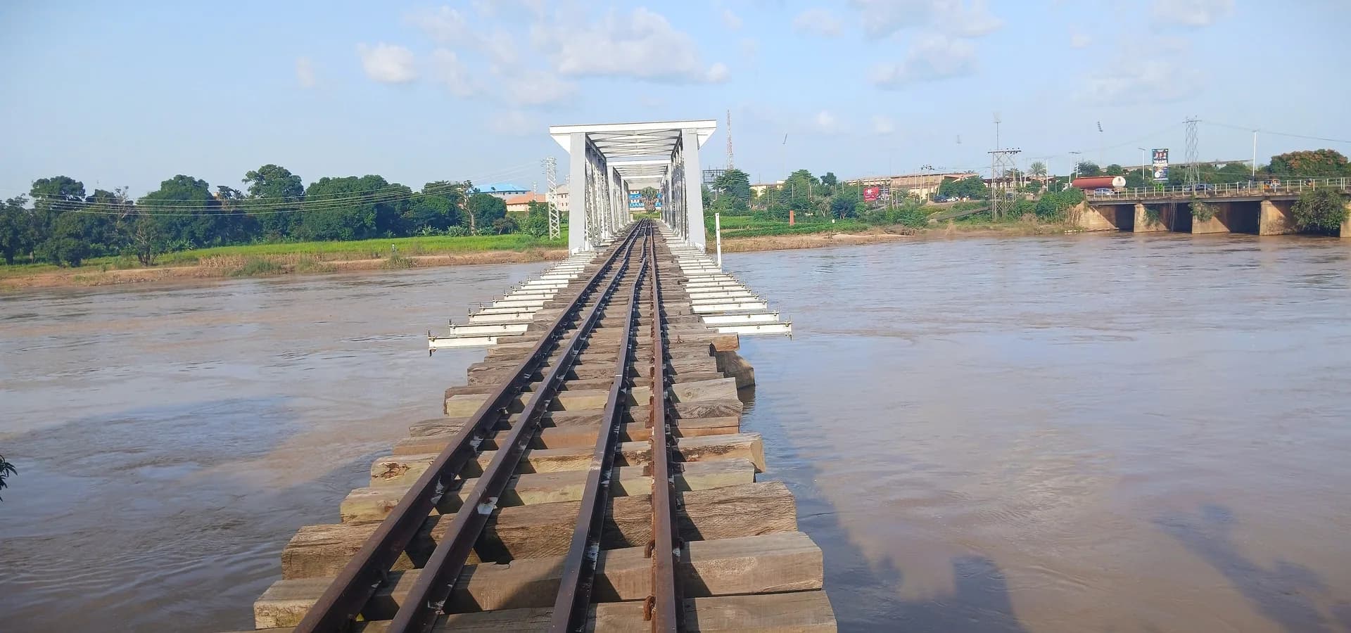 Rail tracks leading across a colonial-era bridge over the Kaduna River, with trees and city buildings on the far bank