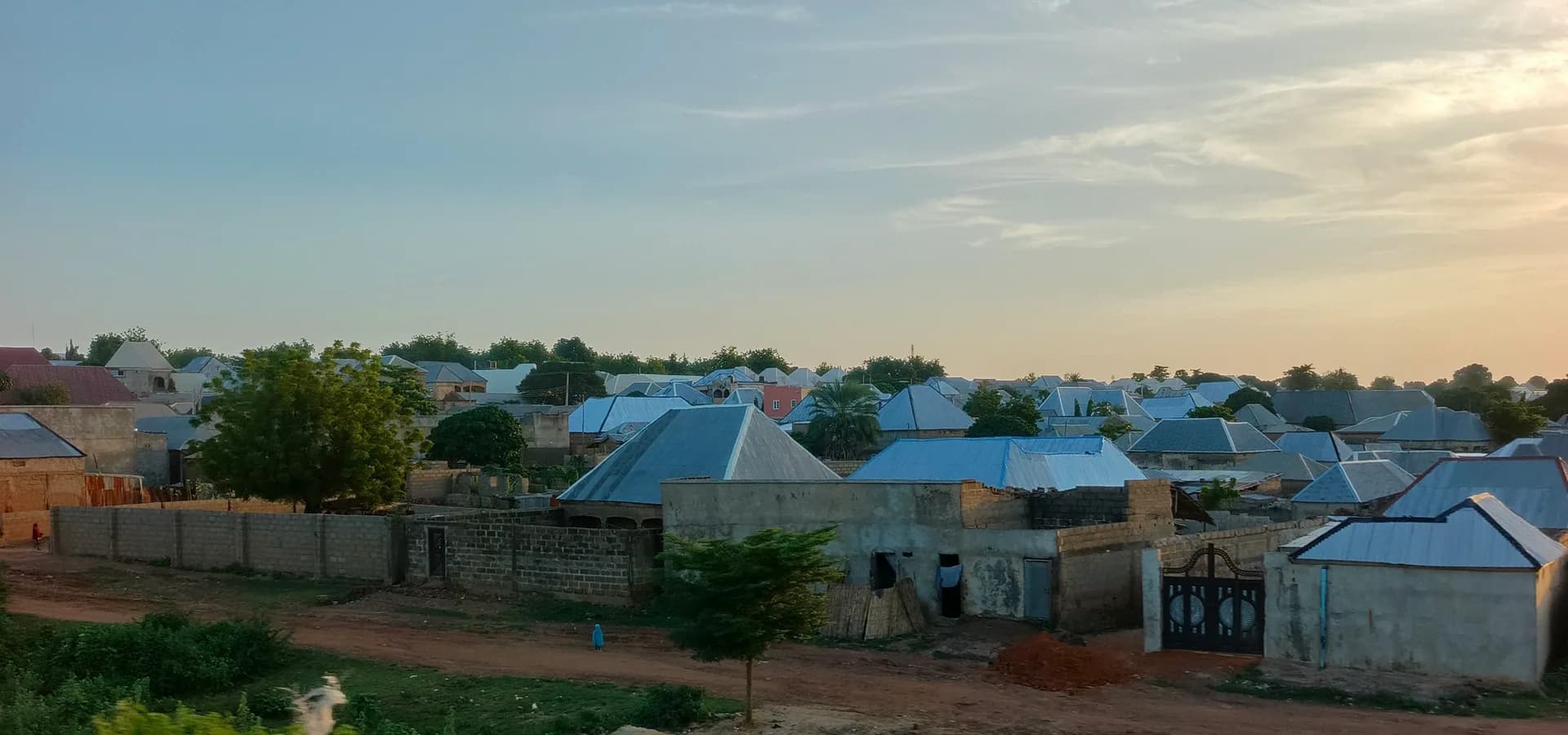 Blechdächer und Bäume eines Viertels in Sokoto unter blassem Abendhimmel