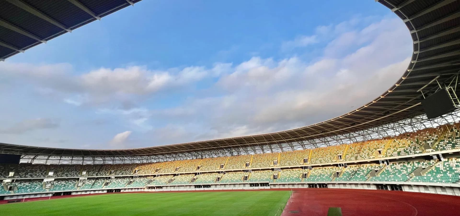 Arquibancadas curvas, gramado verde e pista vermelha do estádio Nest of Champions, em Uyo, sob um céu azul