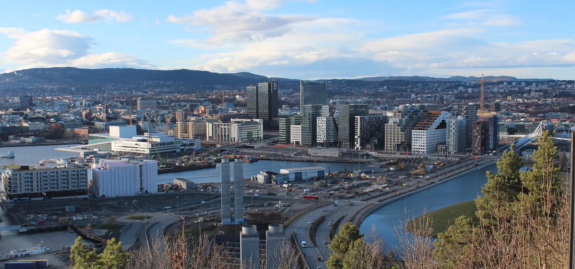Blick vom Hügel Ekeberg über Oslo mit Opernhaus, Türmen von Bjørvika und bewaldeten Hügeln unter blauem Himmel