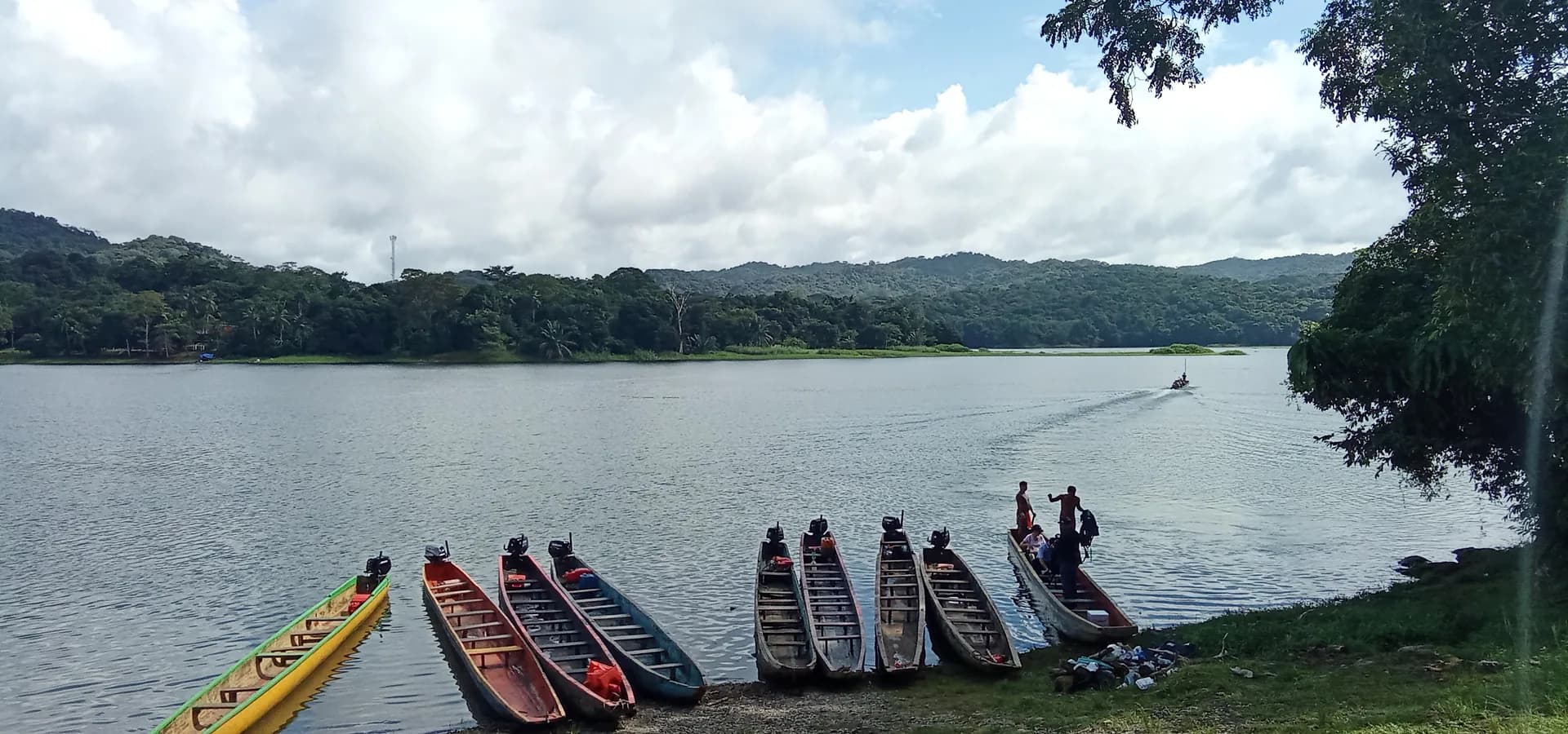 Coloridas piraguas alineadas en la orilla del lago Alajuela, con colinas boscosas y nubes blancas al fondo