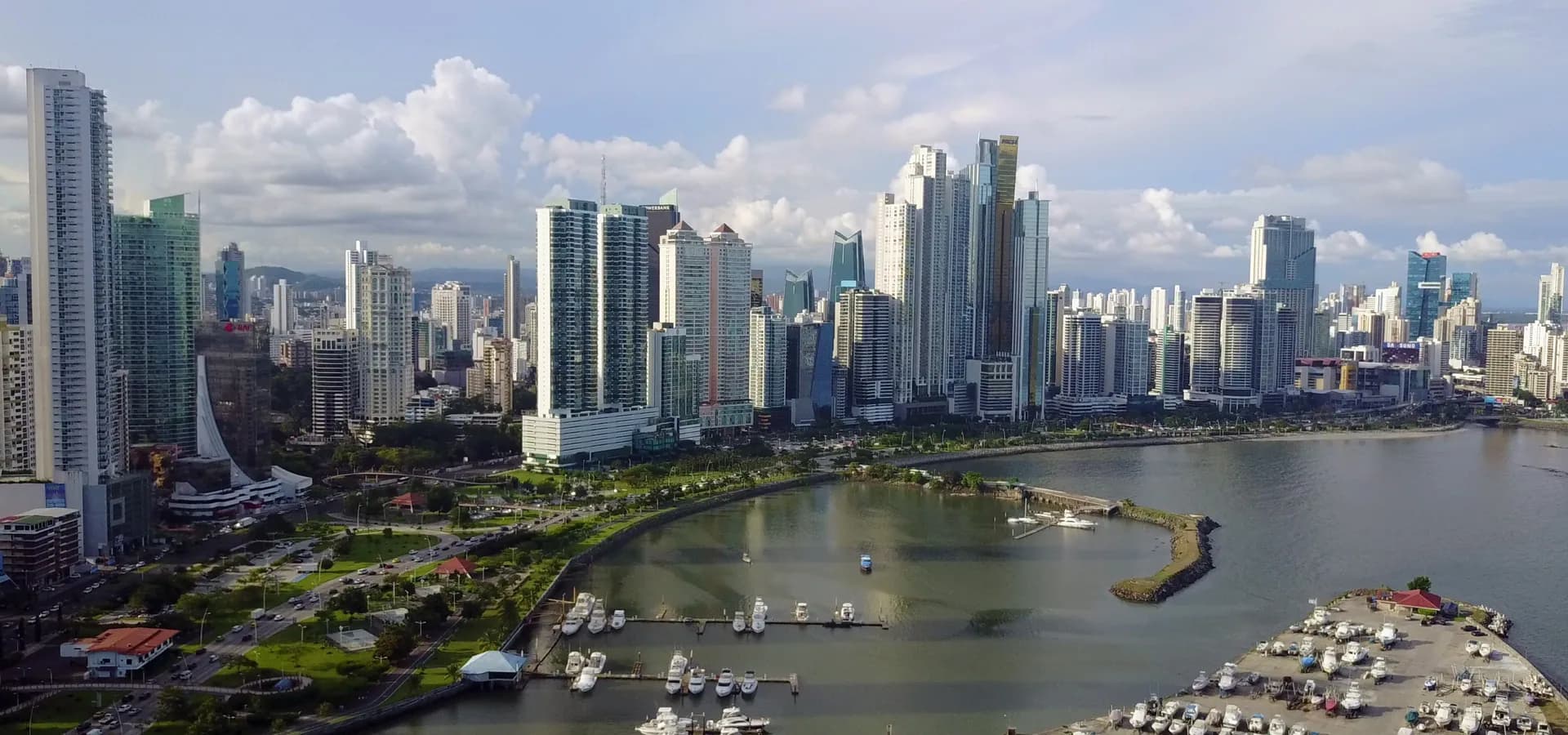 El skyline de rascacielos de Ciudad de Panamá tras una bahía tranquila y una marina llena de barcos bajo nubes blancas.