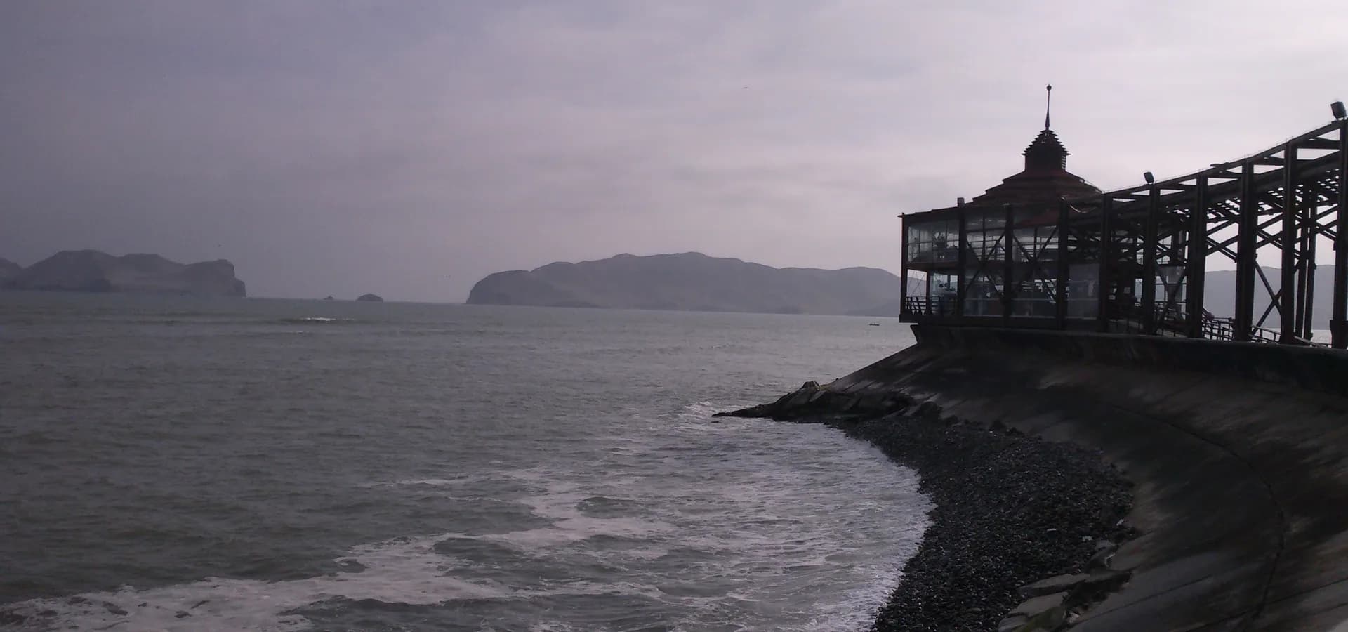 Pazifikwellen brechen am Kieselufer von La Punta, rechts ein hölzerner Pier-Pavillon, am Horizont die Insel San Lorenzo.