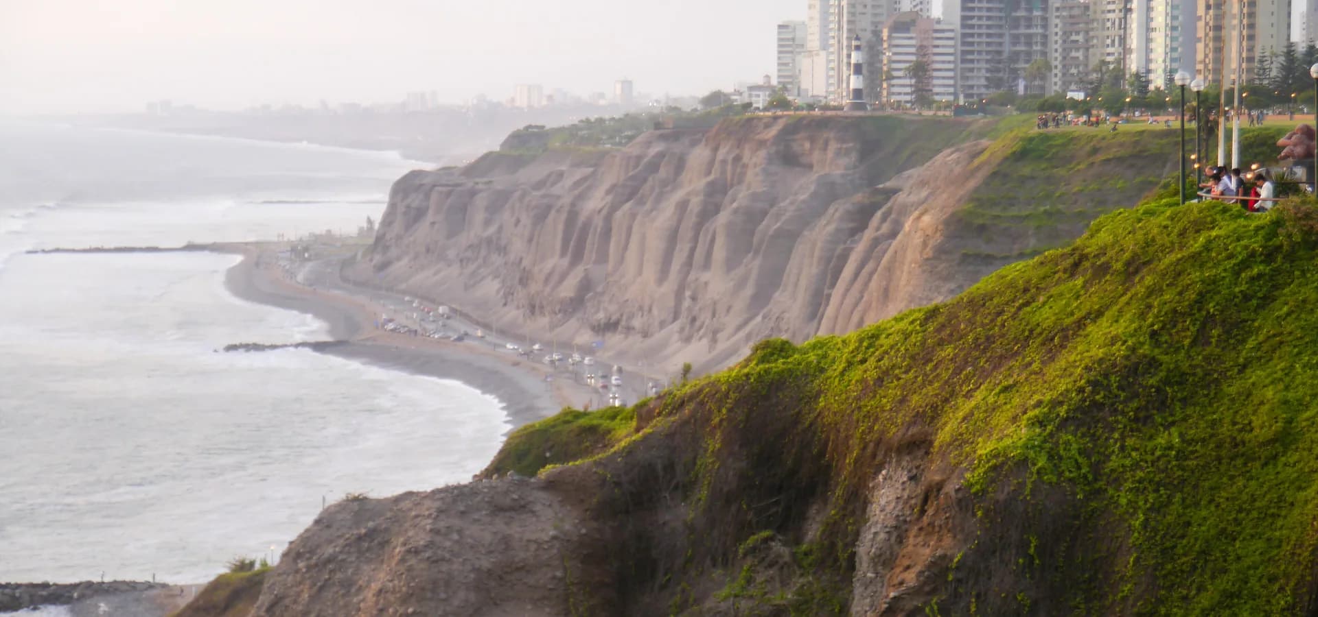 Green and brown cliffs of the Costa Verde above the Pacific, with Miraflores towers and a lighthouse on top.