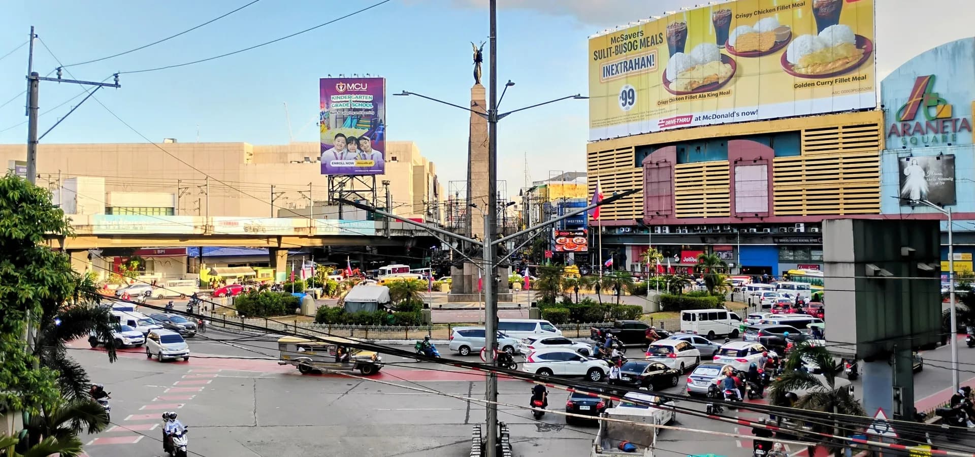 Verkehr kreist um das Monumento in Caloocan, der Obelisk des Bonifacio-Denkmals ragt zwischen Malls unter blauem Himmel auf