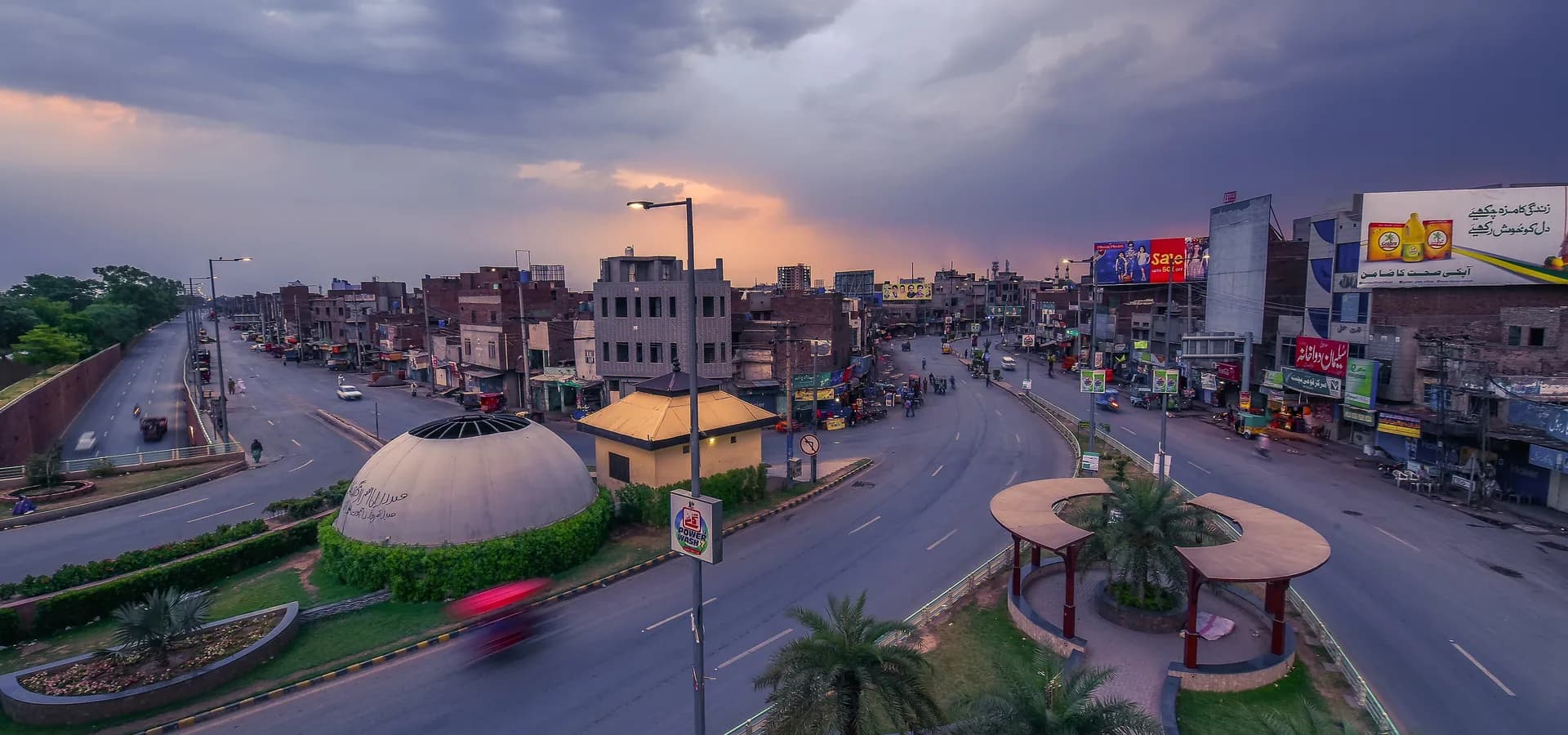 Elevated view of Jhal Chowk in Faisalabad, palm-lined roundabout and busy streets under dramatic evening storm clouds