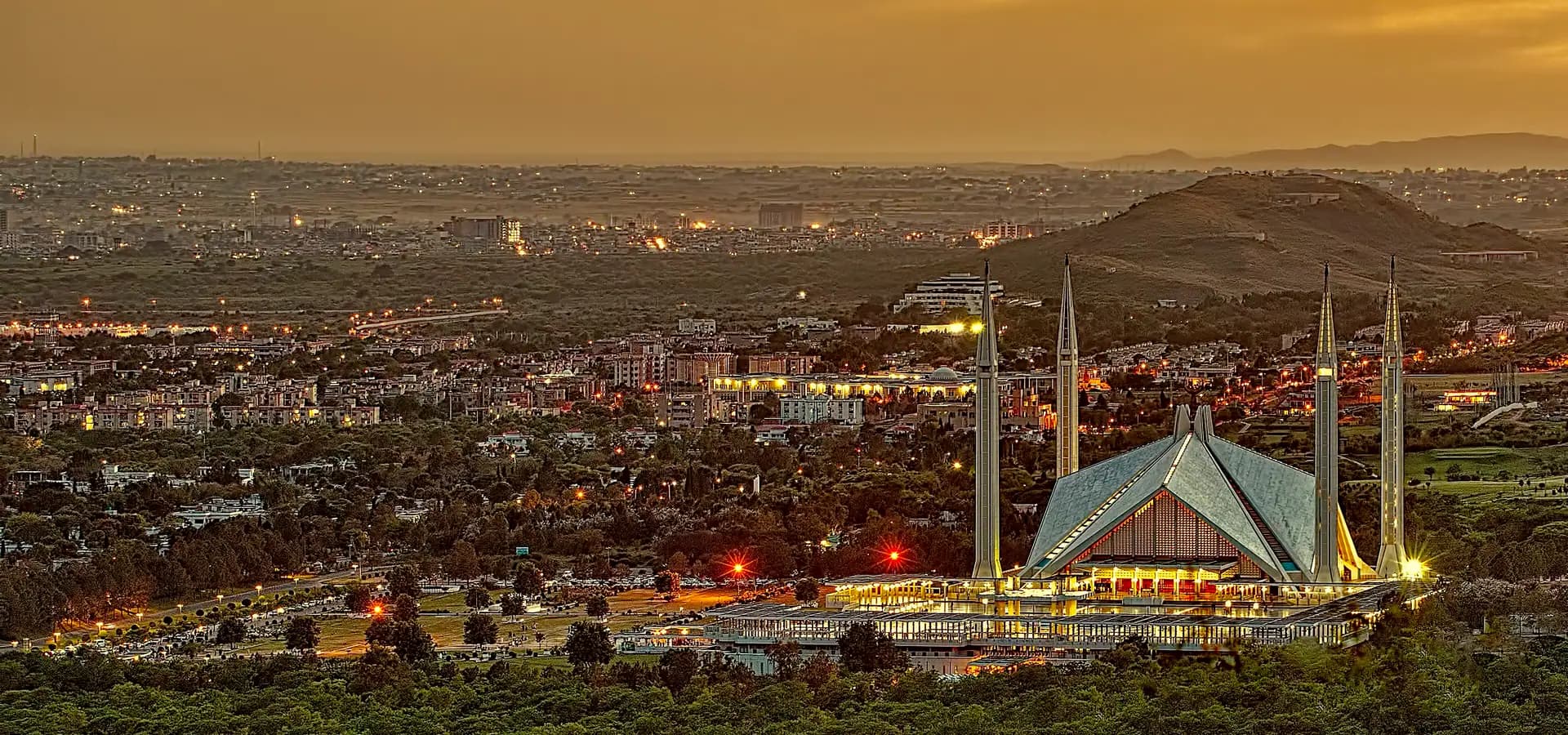 Islamabad under a golden dusk sky, with the lit Faisal Mosque and its minarets rising above green trees