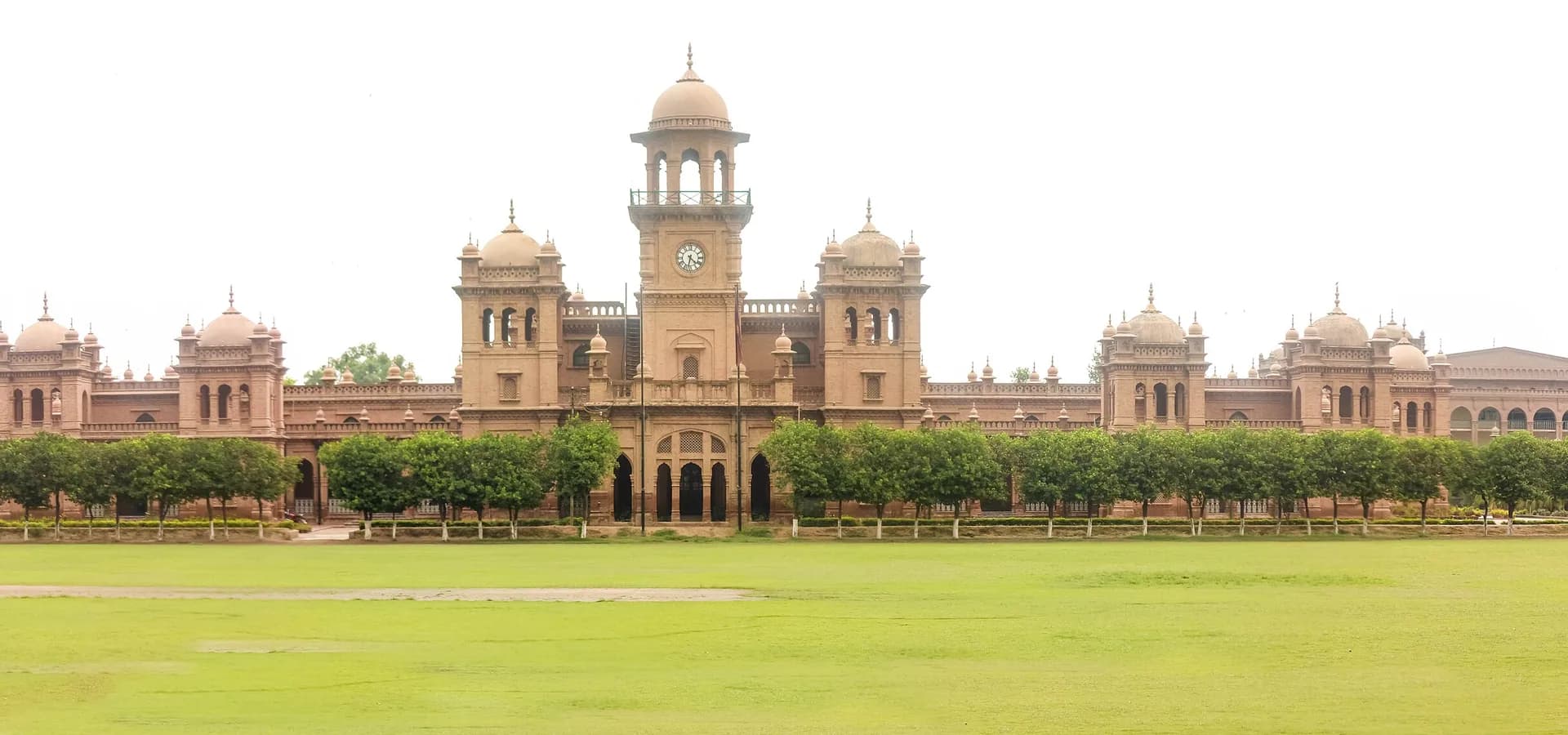 Weite Ansicht der roten Backsteinkuppeln und des Uhrturms des Islamia College Peshawar hinter Bäumen und einem breiten grünen Rasen