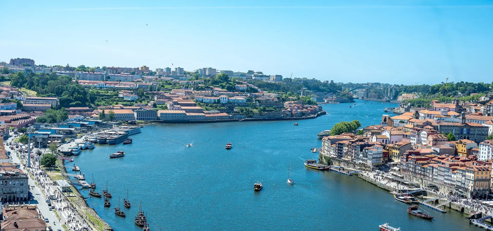 O largo rio Douro no Porto com barcos rabelo, os cais de Gaia à esquerda e as casas da Ribeira à direita sob céu azul