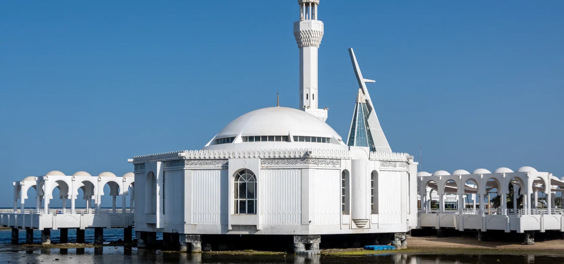 Die weiße Al-Rahmah-Moschee auf Stelzen über ruhigem Wasser des Roten Meeres in Dschidda, ihr Minarett vor klarblauem Himmel