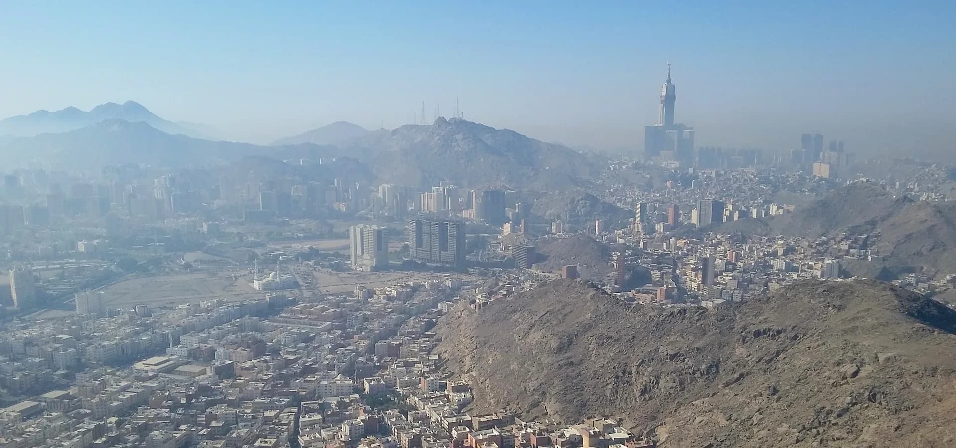 Meca vista do alto de uma colina, fileiras de prédios entre colinas rochosas e as Torres do Relógio se erguendo na bruma