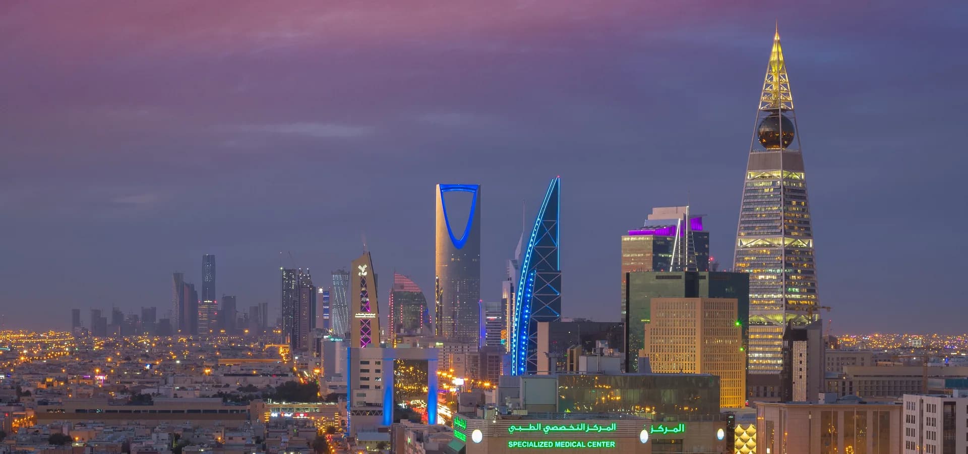 Horizonte de Riade ao anoitecer, com o arco do Kingdom Centre e a Torre Al Faisaliah coroada por um globo sob um céu violeta