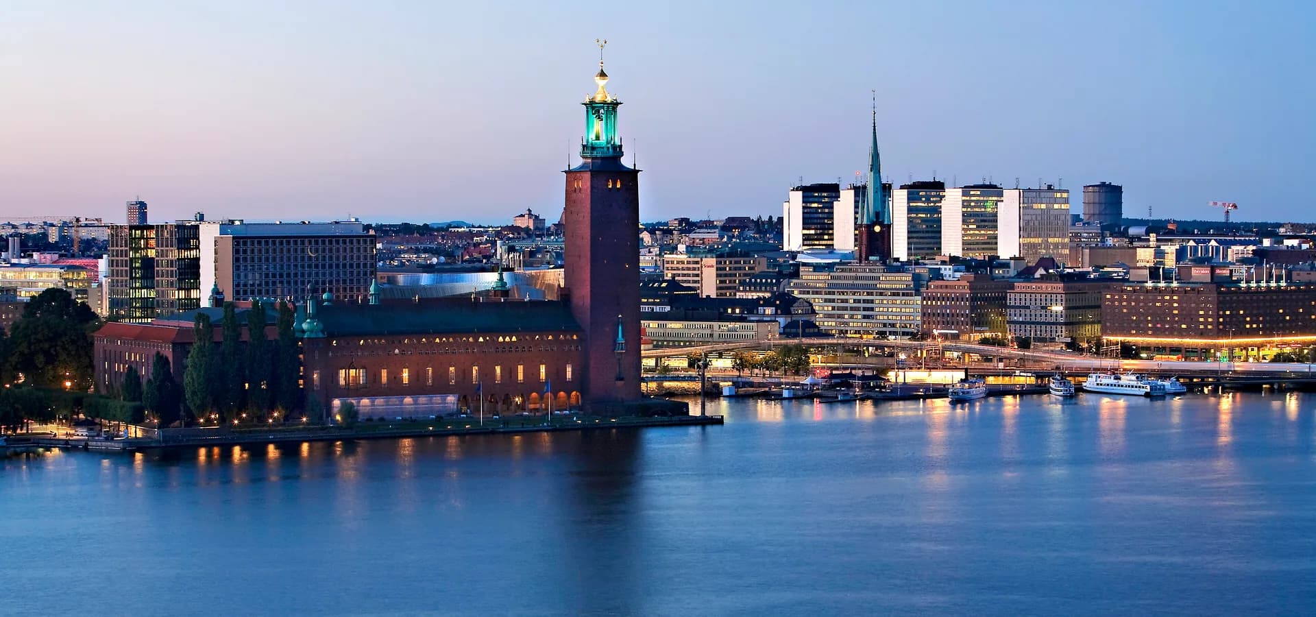 Stockholm City Hall tower lit at dusk across the calm water of Riddarfjärden, with the city skyline behind