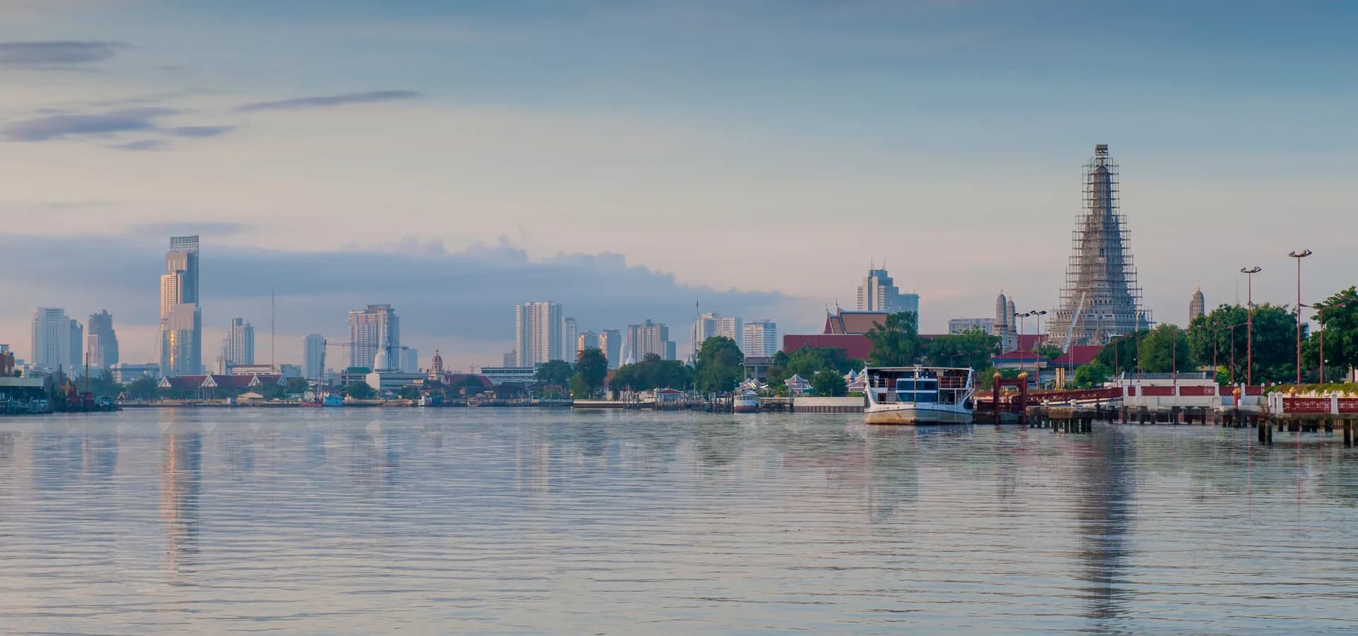 Stilles, pastellfarbenes Wasser des Chao Phraya bei Morgendämmerung, Bangkoks Hochhäuser am Horizont und Wat Arun rechts