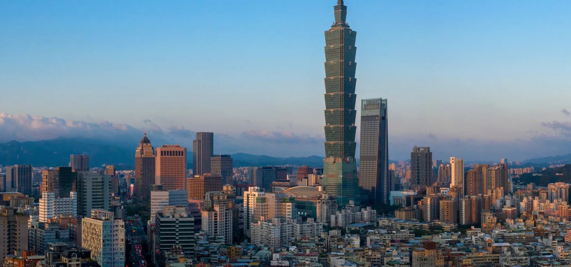 Taipei 101 glowing in late sun over a dense city, with green mountains and a soft blue sky beyond