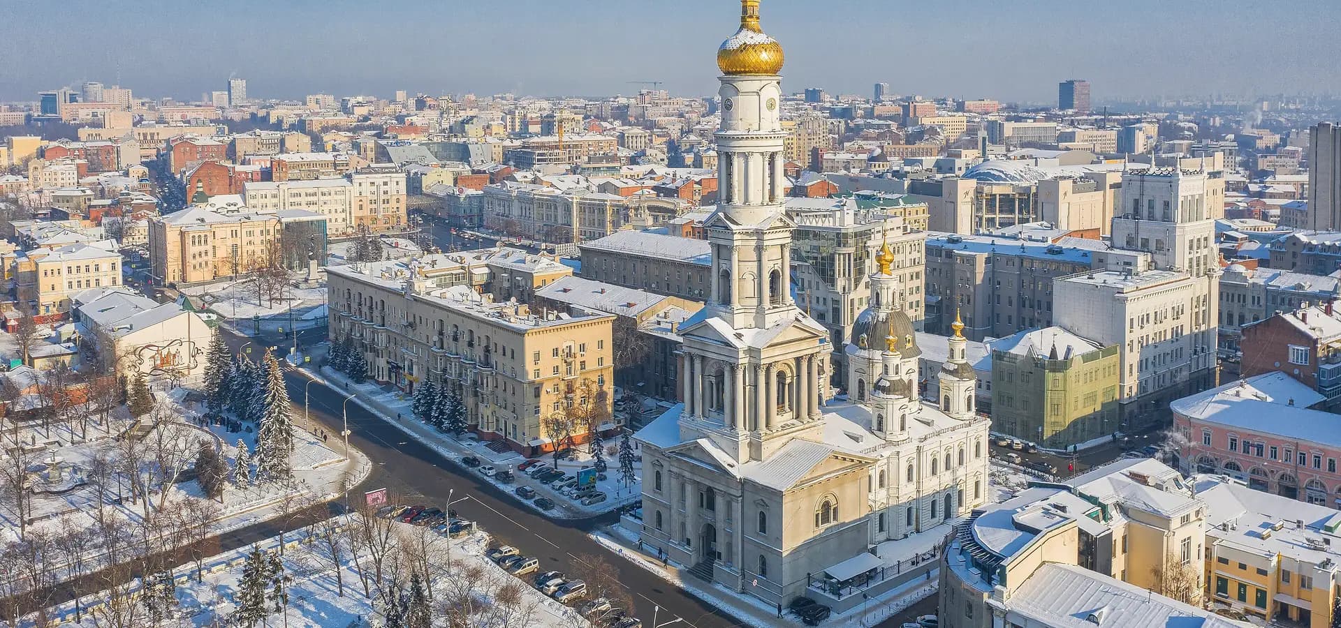 Vista aérea de inverno da torre sineira branca da Catedral da Dormição, com cúpula dourada, sobre telhados nevados do centro de Kharkiv