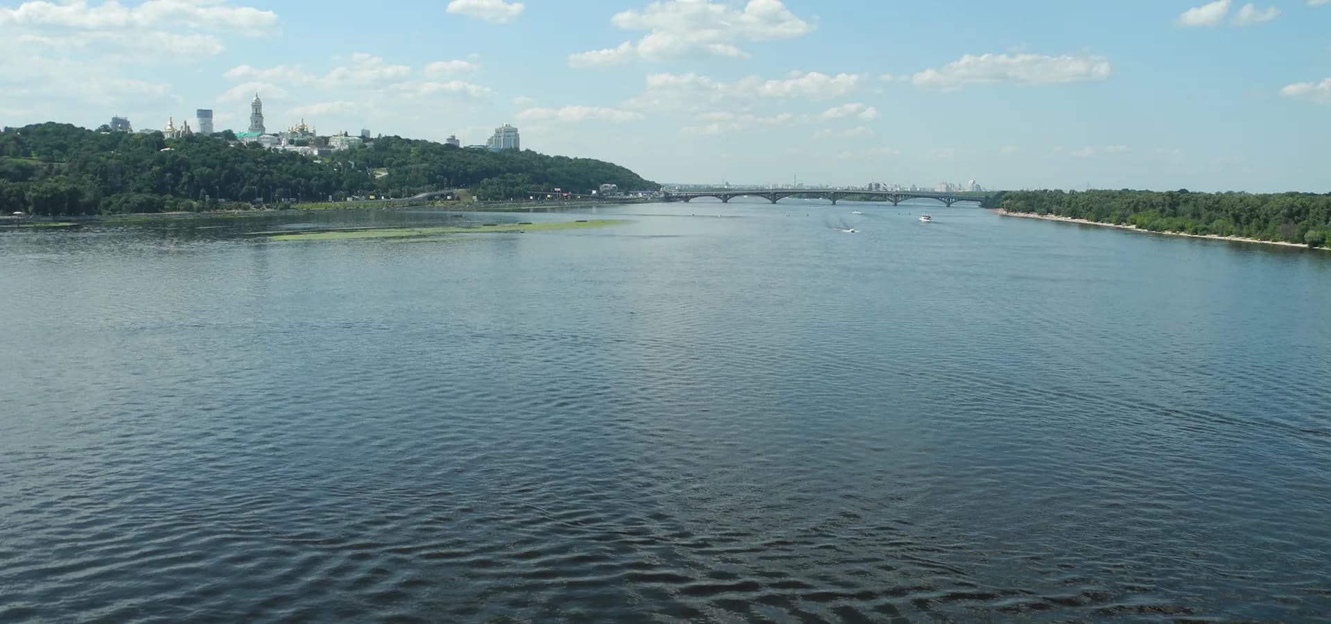 Der breite Dnipro unter dem Sommerhimmel, mit grünen Hügeln und den goldenen Kuppeln des Höhlenklosters auf den Hügeln links
