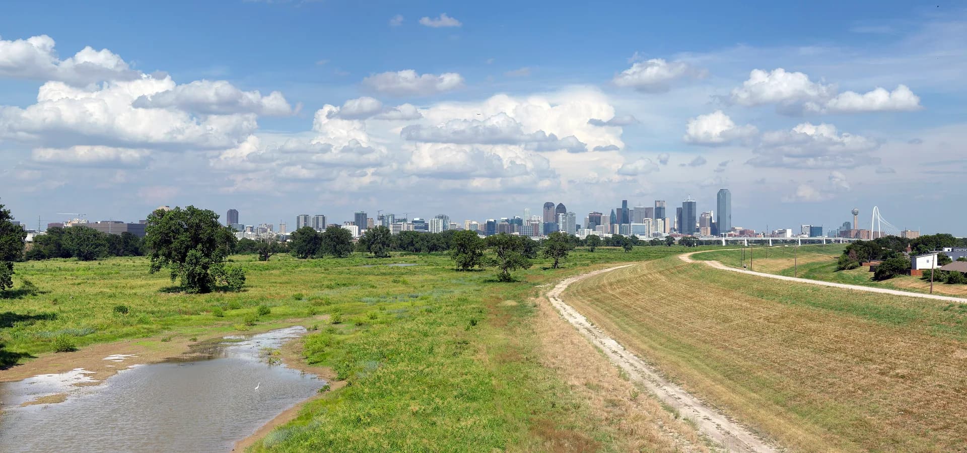 Dallas skyline on the horizon beyond the green Trinity River floodplain, a levee path and white clouds