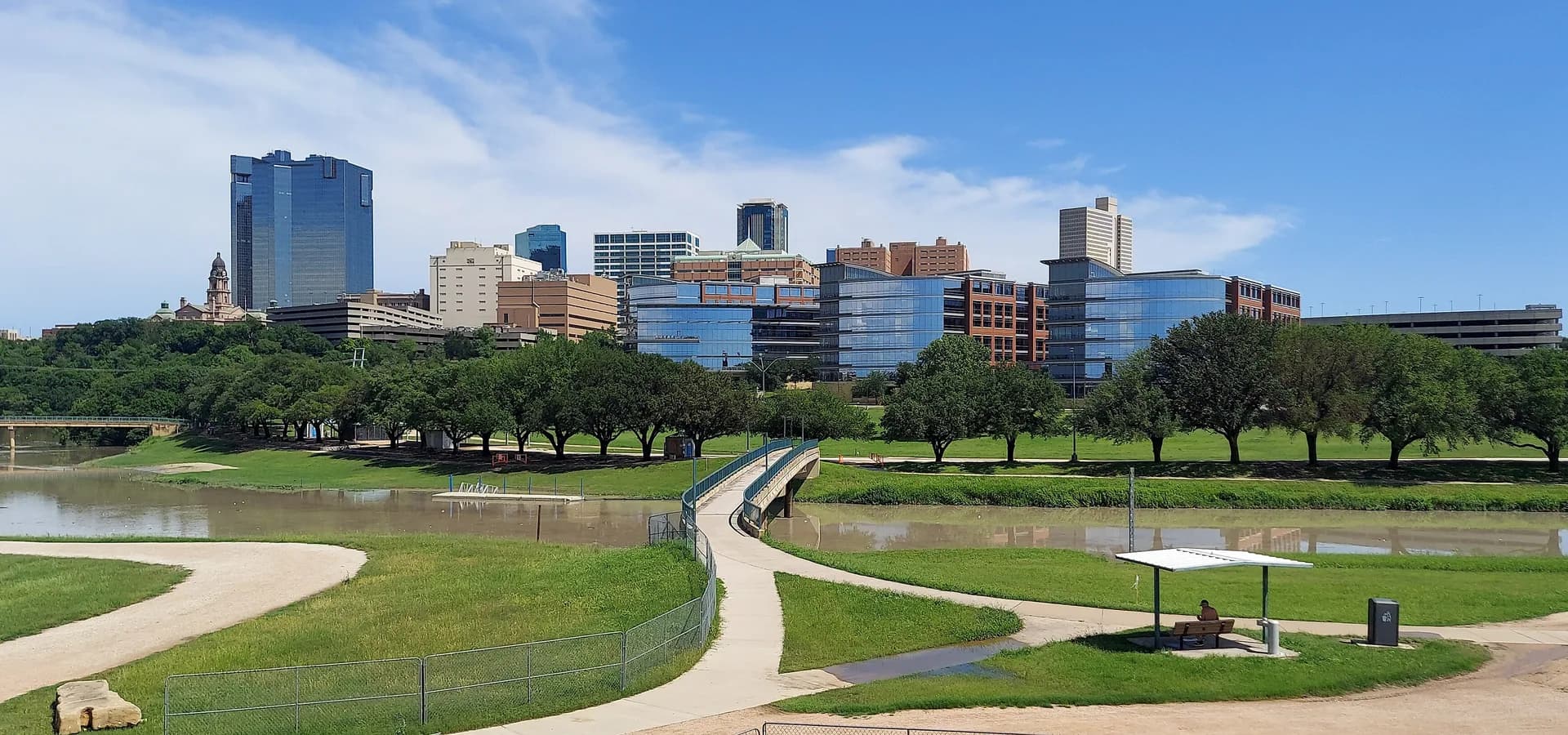 Torres do centro de Fort Worth surgindo atrás de árvores e do rio Trinity sob um céu azul de verão