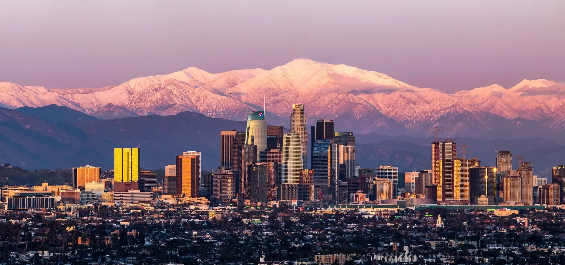 Prédios do centro de Los Angeles iluminados ao entardecer diante das montanhas San Gabriel nevadas e do Mount Baldy