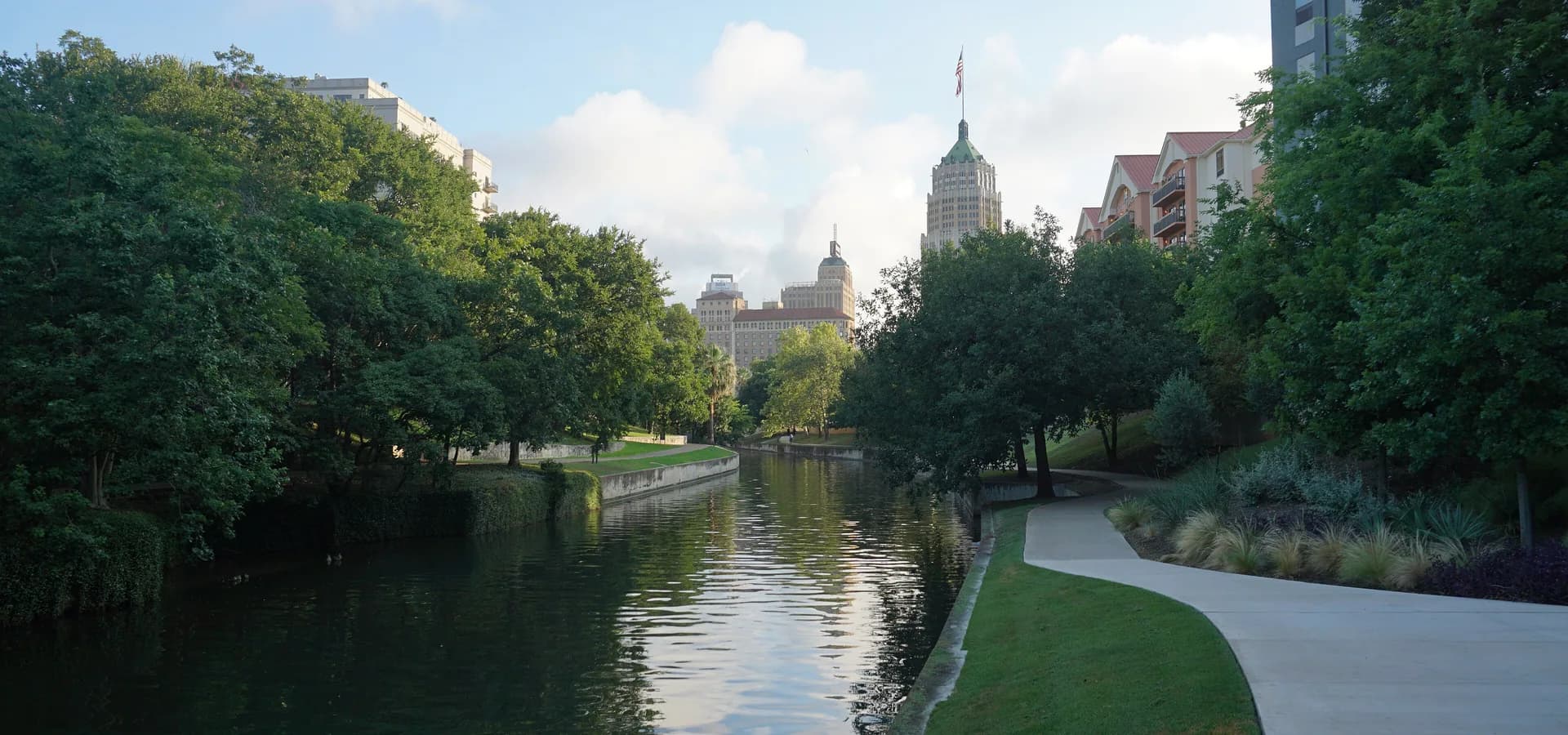 The calm San Antonio River framed by trees and a curving path, with the Tower Life Building rising downtown