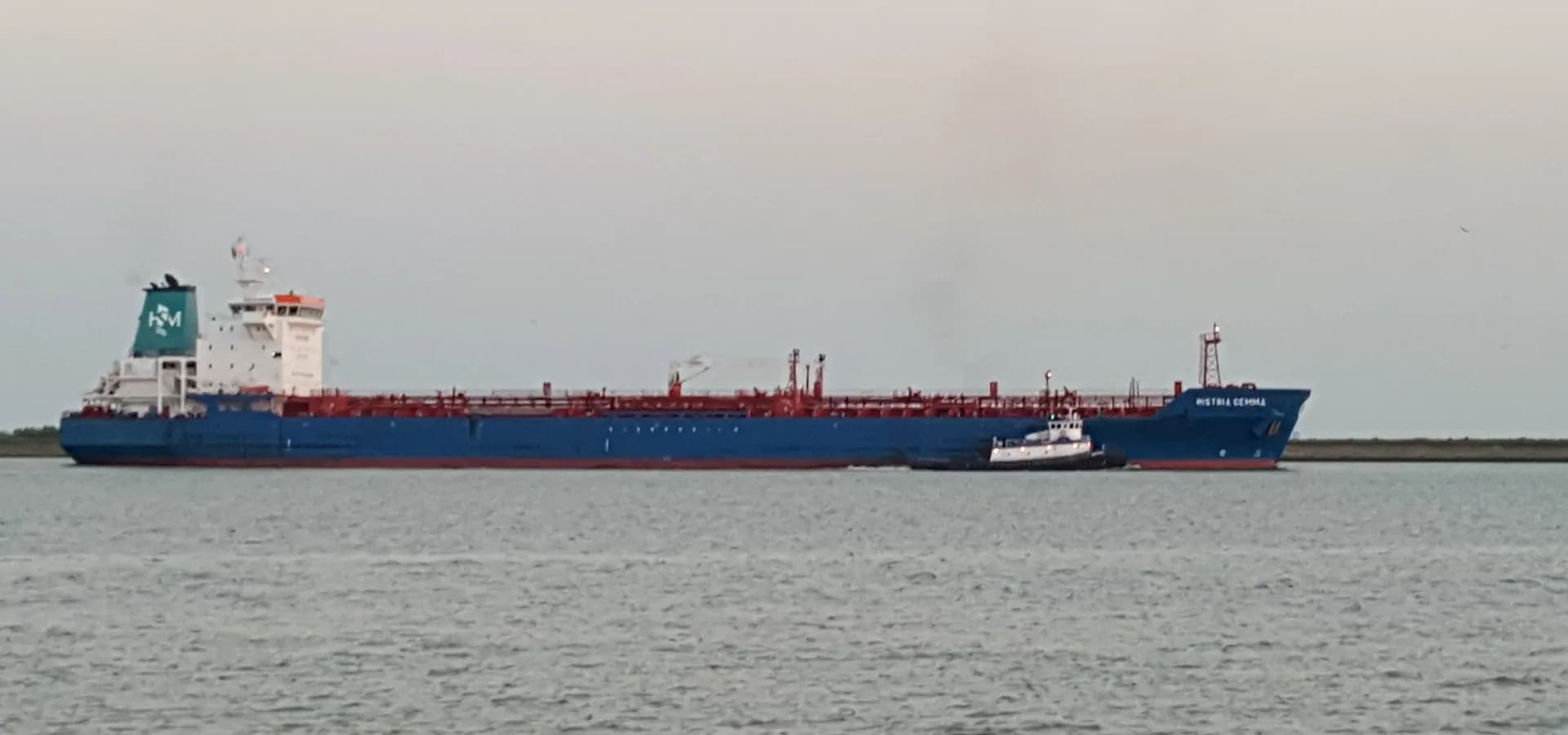Un petrolero azul y rojo con un remolcador deslizándose por el canal junto al Texas City Dike bajo un cielo pálido