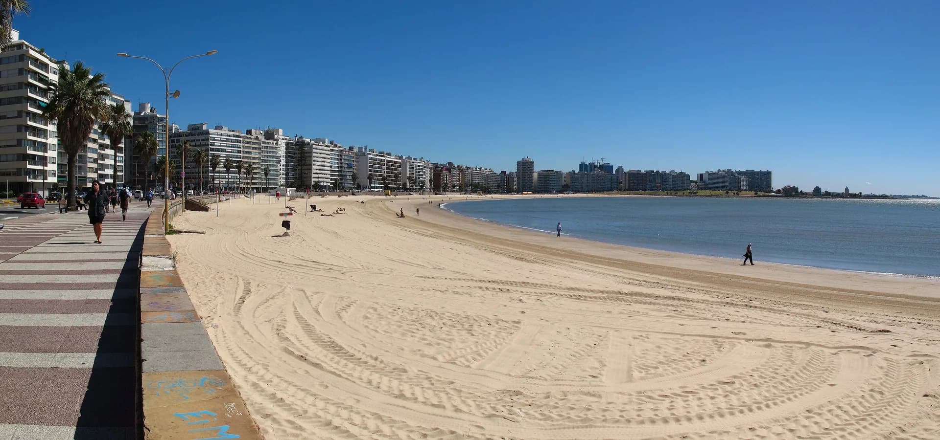 Die weite Sandbucht des Strandes Pocitos in Montevideo mit der Rambla und weißen Wohntürmen unter blauem Himmel