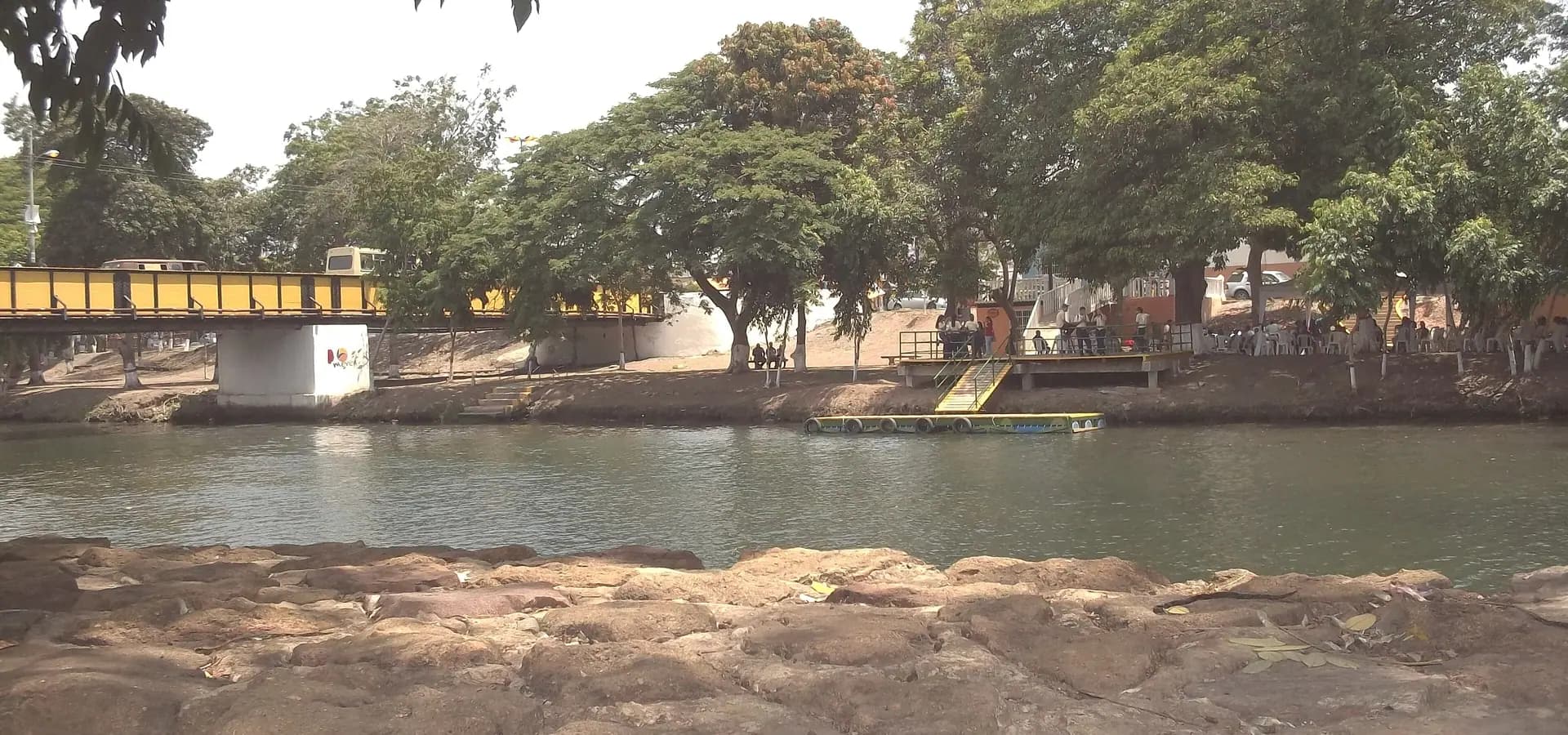The calm green Neverí River in Barcelona, Venezuela, with a yellow footbridge and shade trees along the bank
