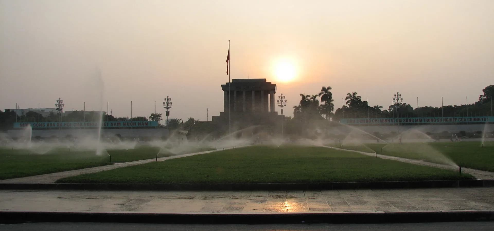 Wide green lawns of Ba Đình Square with sprinklers running and the granite mausoleum under a hazy sunset