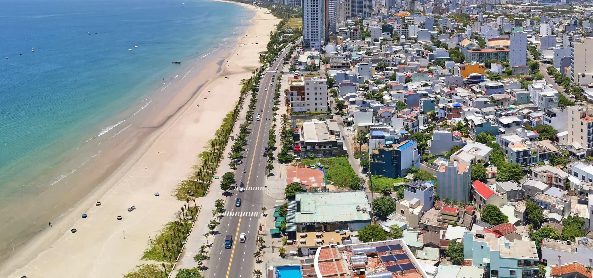 Luftbild eines langen weißen Strandes mit türkisfarbenem Meer links und den Türmen der Strandpromenade von Da Nang rechts