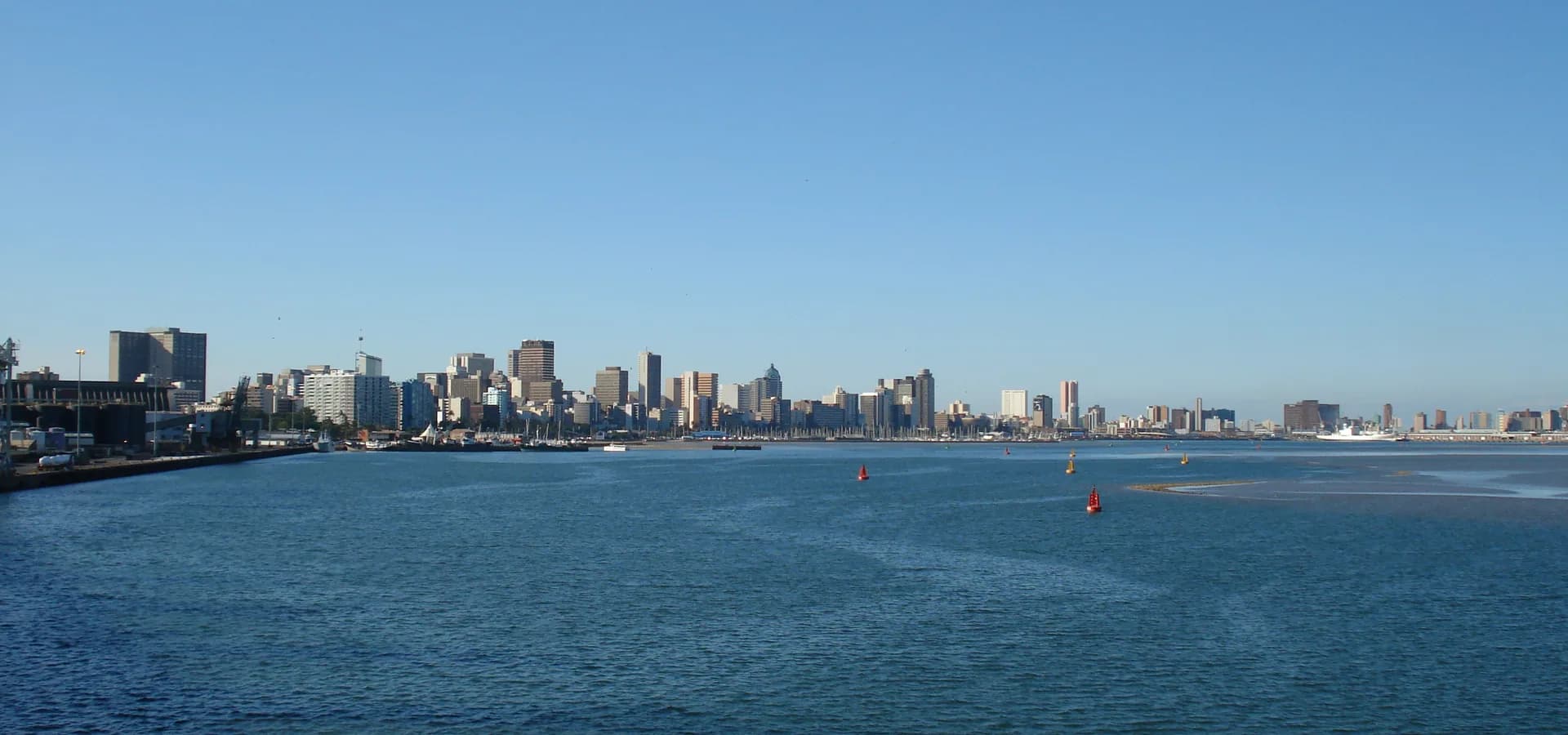 Die Skyline der Innenstadt von Durban zieht sich unter klarem blauem Himmel am Hafen entlang, davor ruhiges Wasser