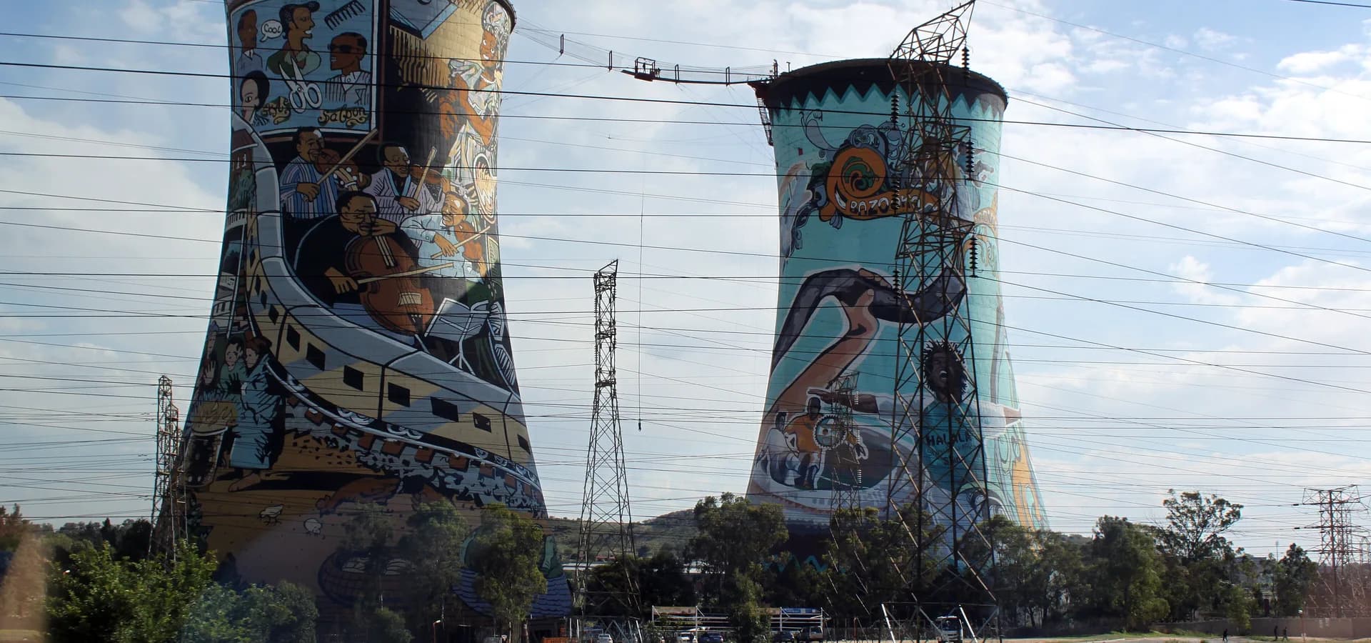 Dos torres de refrigeración pintadas en Soweto, una con un mural de músicos de jazz, se alzan sobre un terreno verde abierto
