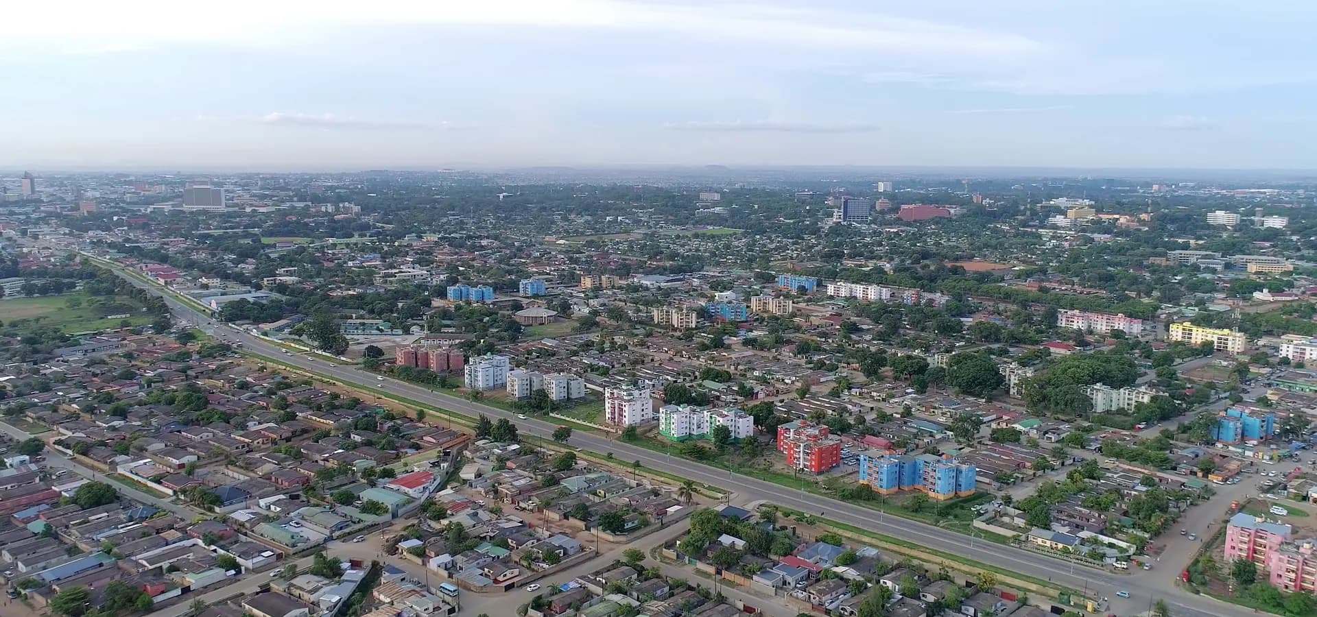 Weite Luftaufnahme über grüne Vororte von Lusaka, Wohnblöcke und eine lange Schnellstraße Richtung ferner Türme