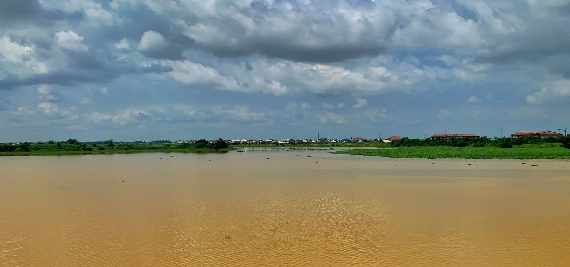येनागोआ में बड़े सफेद बादलों के नीचे चौड़ी नदी, हरे किनारे और दूर किनारे पर छतें