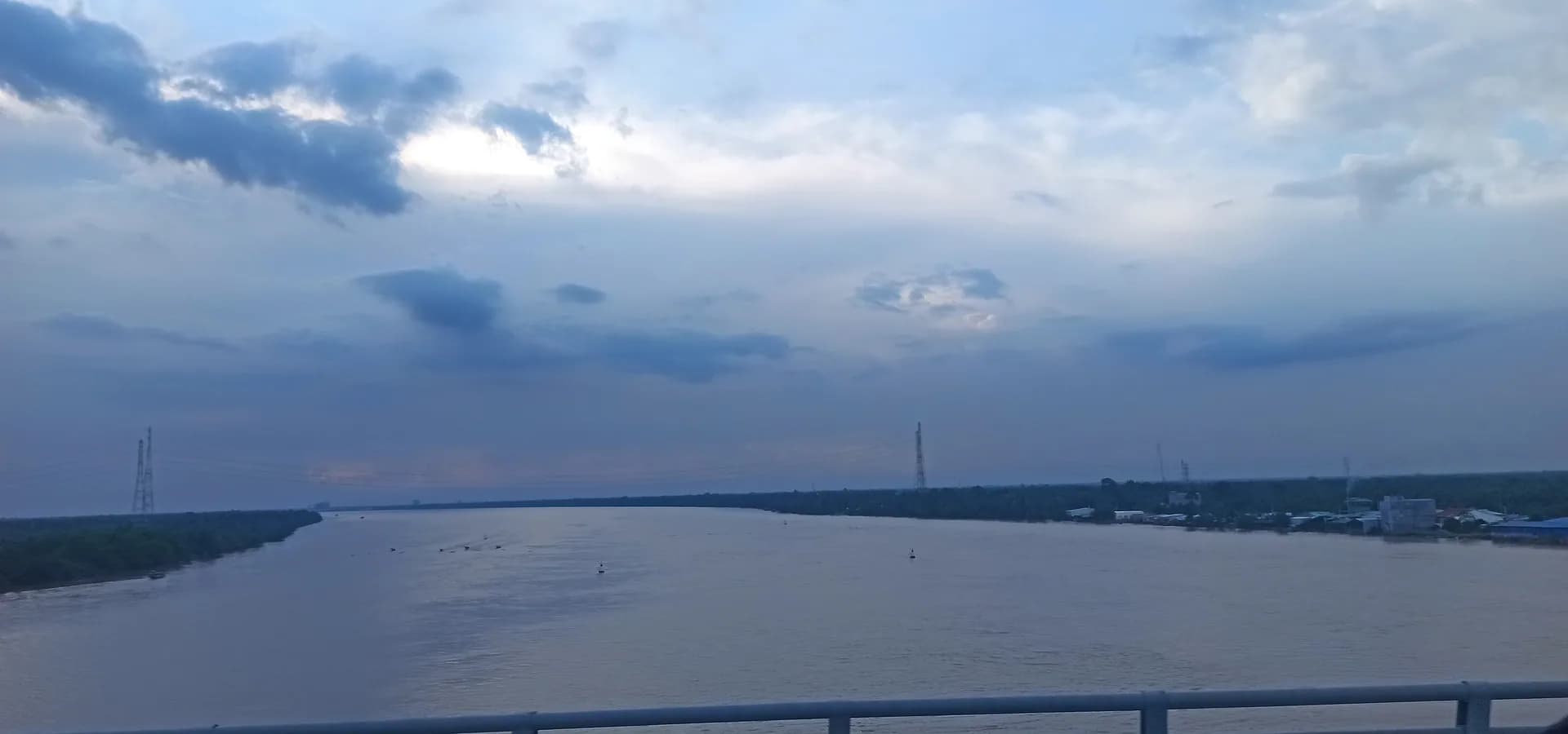 Der breite Hàm Luông von der Brücke aus gesehen, grüne Ufer mit Bäumen unter weichem blauem Wolkenhimmel