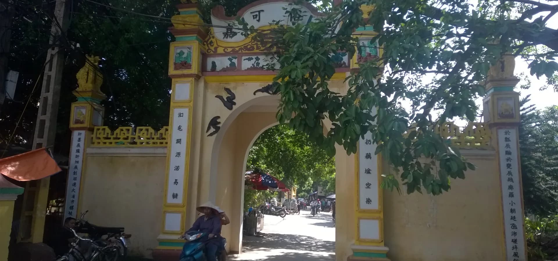Gelbes Dorftor mit Bogen in Hà Đông, chinesische Schriftzeichen auf den Pfeilern, dahinter eine Gasse mit Bäumen